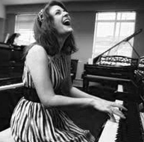 A woman laughing while playing a piano in a room with large windows.