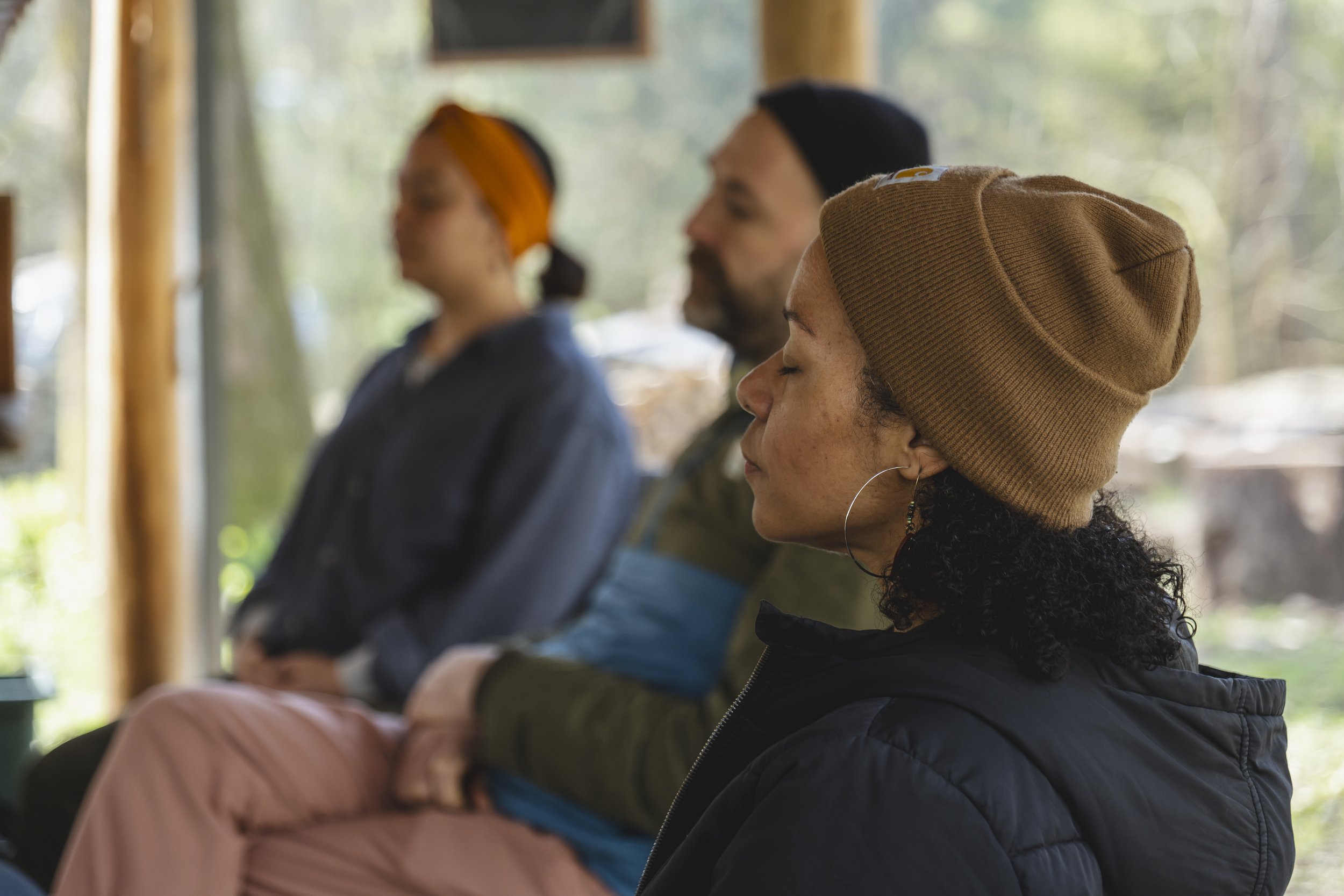 Three people sitting outdoors with eyes closed, suggesting they are meditating or relaxing.