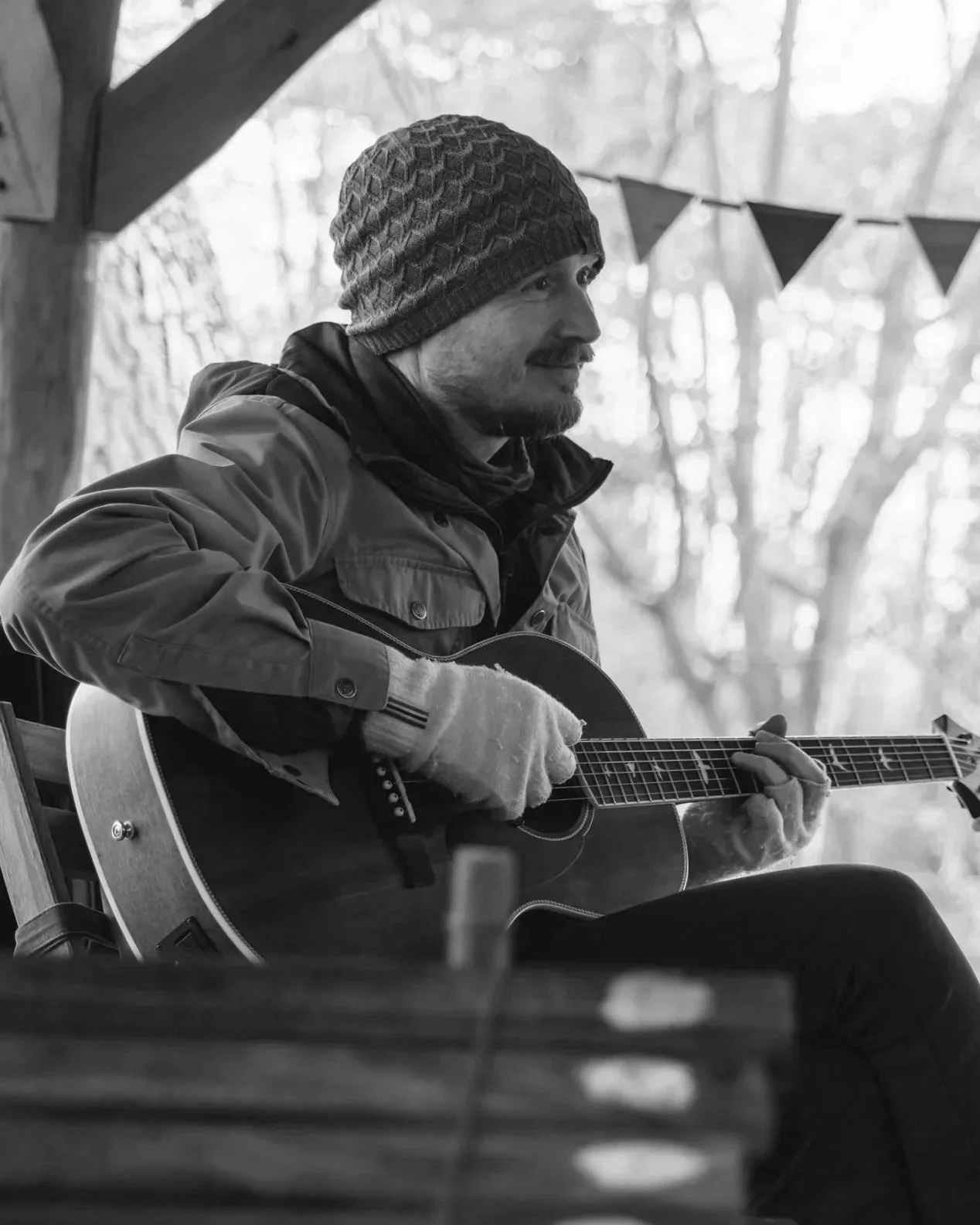 A man in a beanie and jacket playing an acoustic guitar, sitting outdoors in a wooded area with blurry trees in the background.