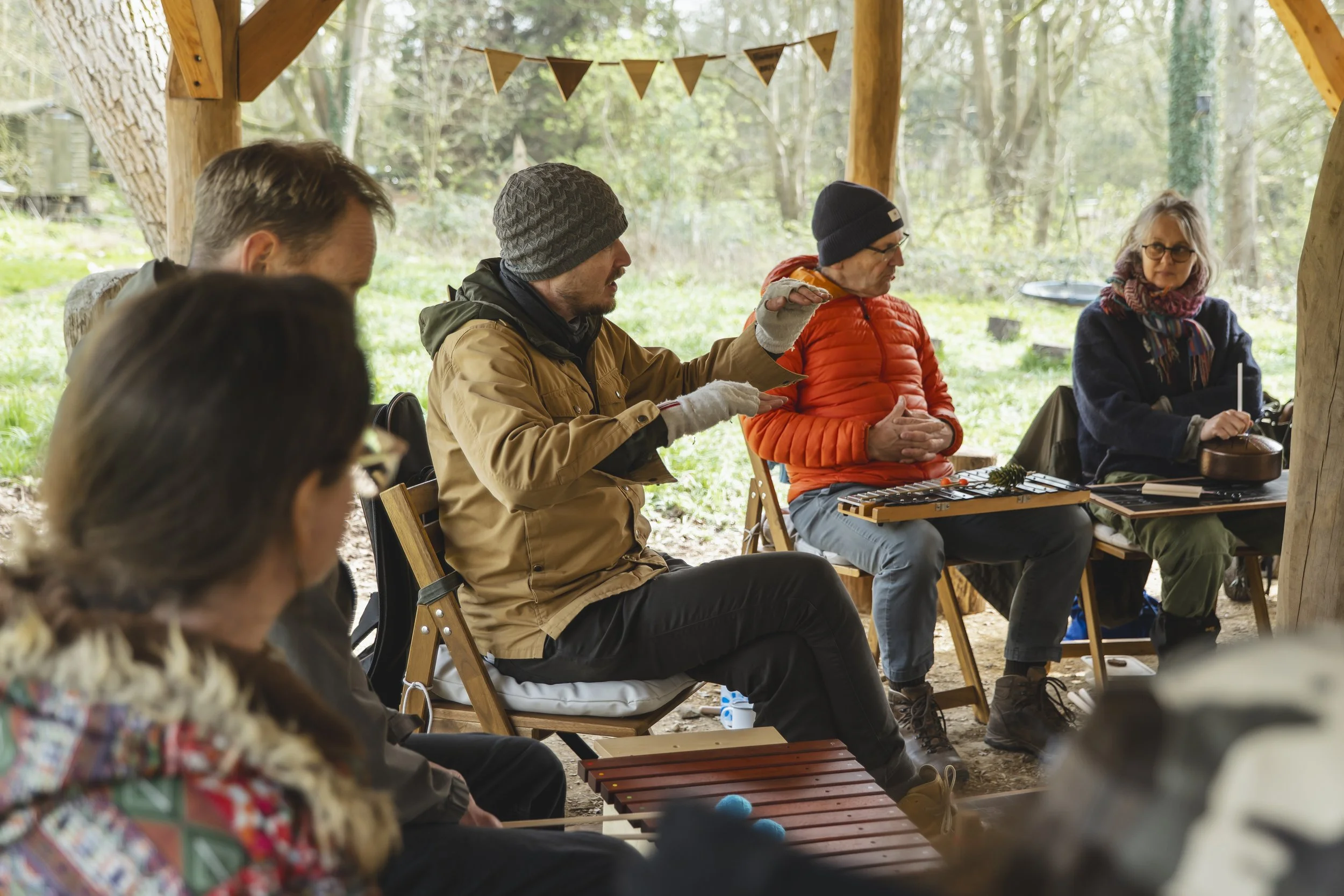 Group of people sitting outdoors under a wooden shelter, engaged in a discussion or workshop, with trees and greenery in the background.