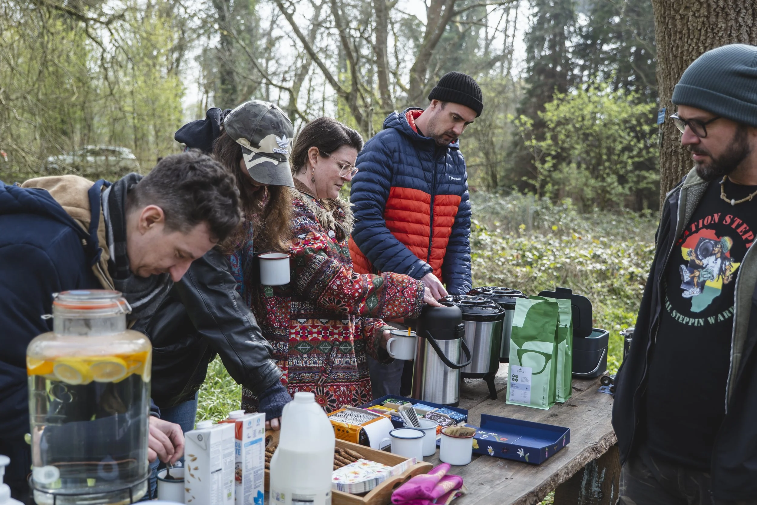 A group of people enjoying an outdoor coffee break at a wooded area with trees and greenery, with various coffee brewing tools and snacks on a wooden table.