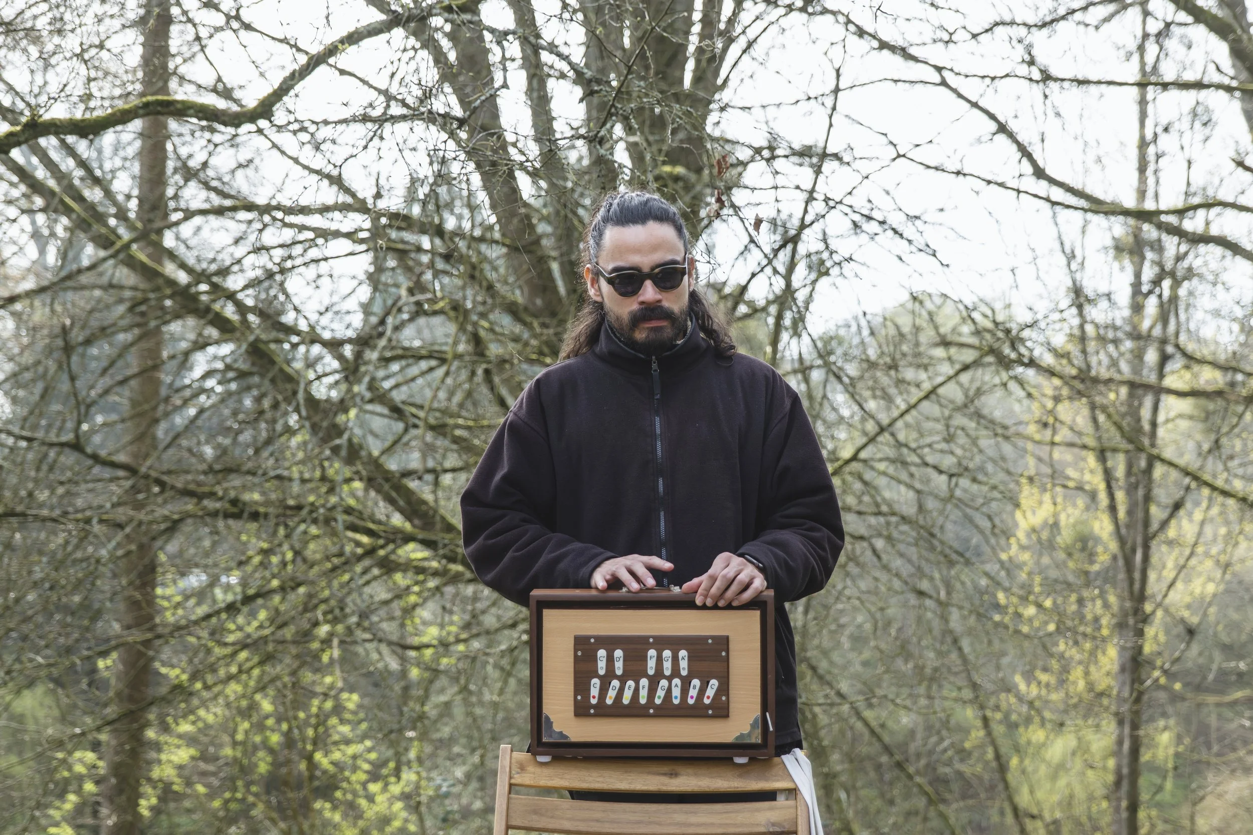 A man with long dark hair and a beard, wearing sunglasses and a black jacket, stands outdoors in a wooded area with leafless trees, holding a shruti box.