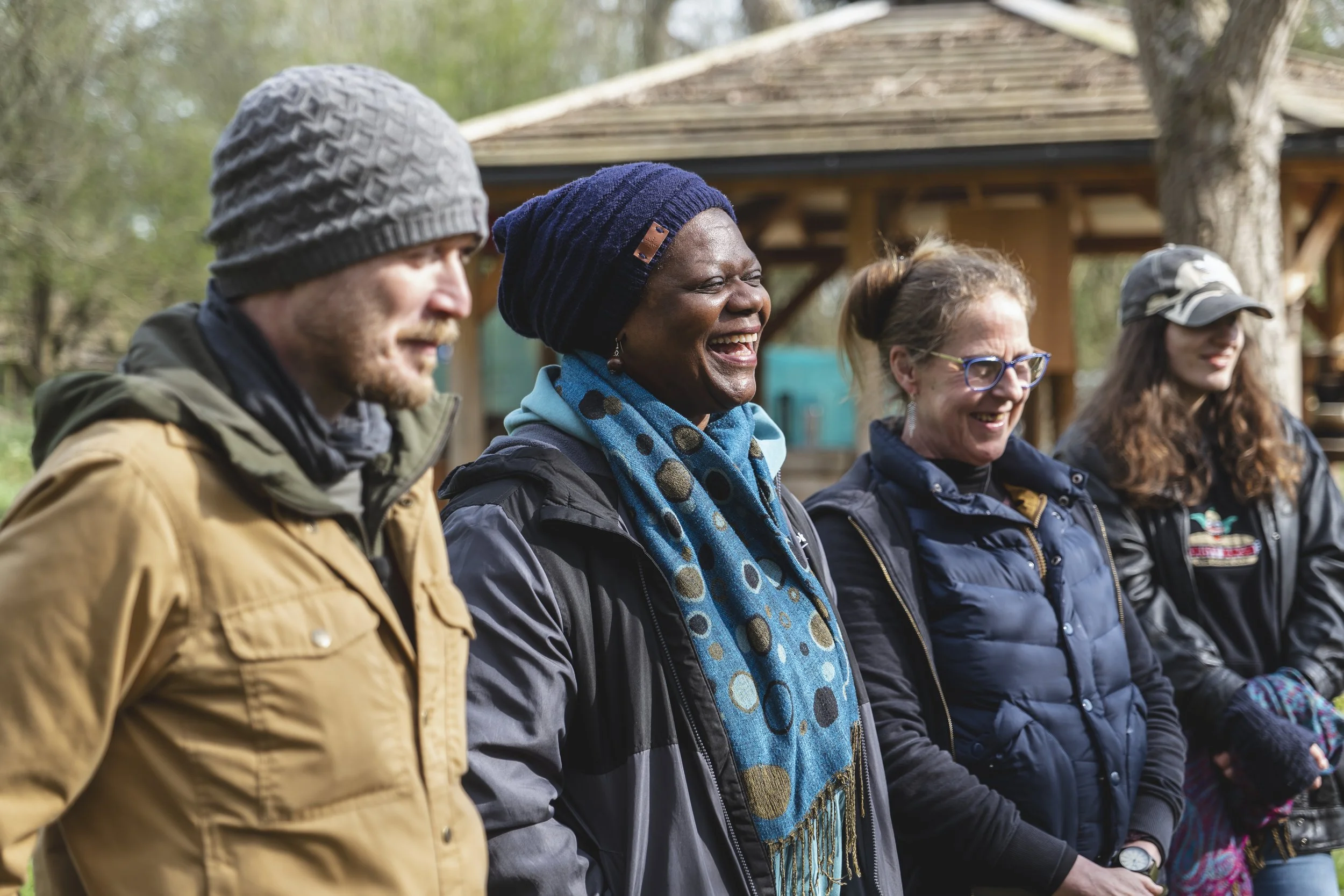 Group of four people outdoors, smiling and laughing, dressed in warm clothes, with trees and a wooden structure in the background.