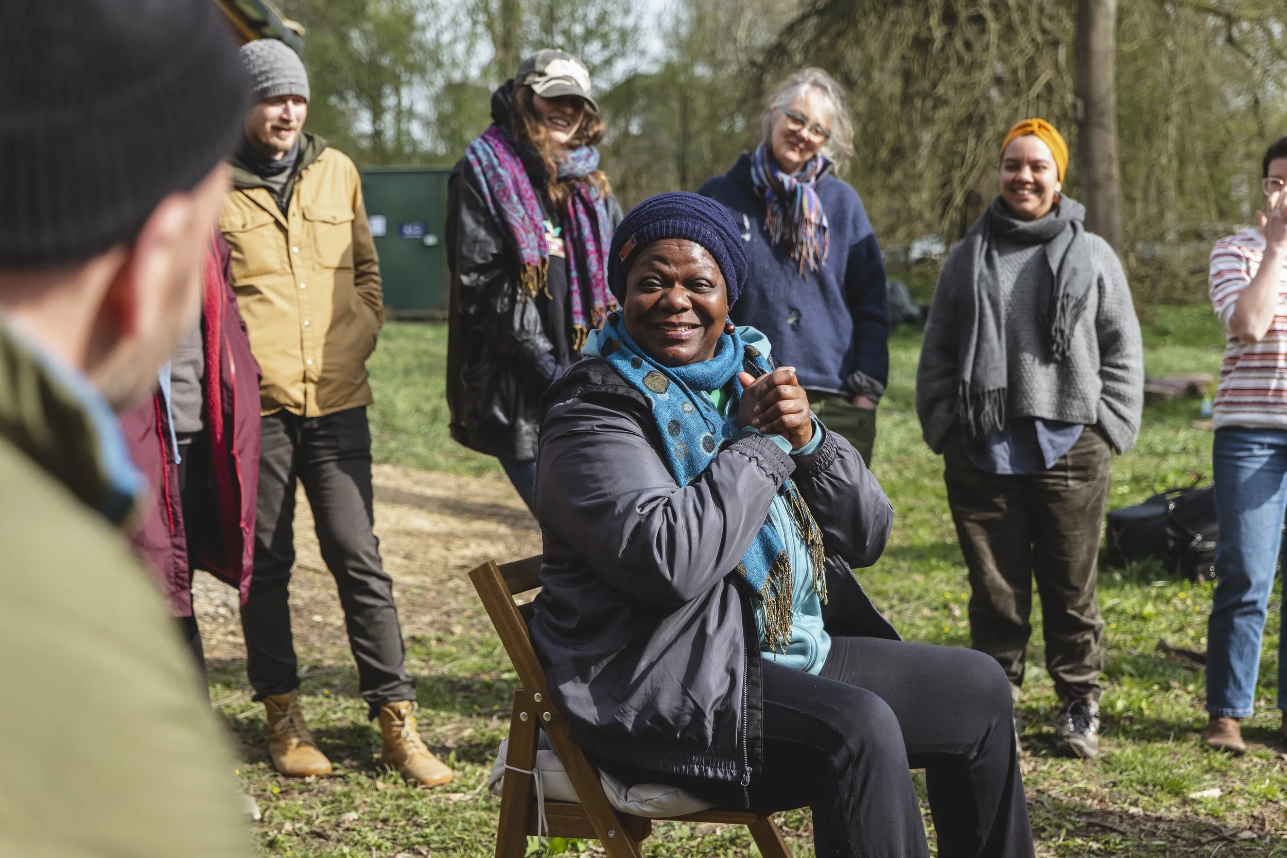 A group of people outdoors, with a woman seated and smiling at the center, surrounded by others standing and smiling.