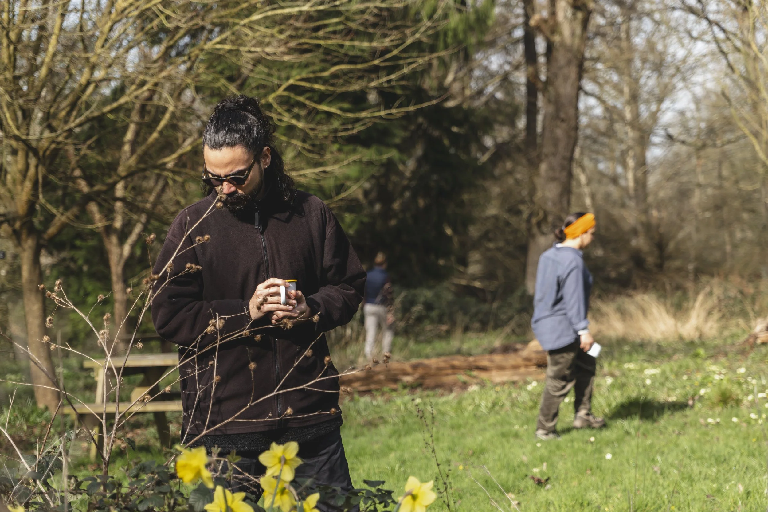 A man with dark hair tied back, wearing sunglasses and a dark jacket, stands outdoors, with yellow flowers and plants in the foreground. Behind him, a woman wearing a gray jacket and orange headscarf is also outdoors in a natural setting with trees a