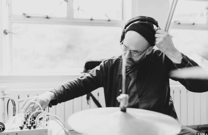 A man wearing headphones and glasses, adjusting his headphones with one hand, sitting at a desk with audio equipment and a cymbal in front of him, in a room with large windows.