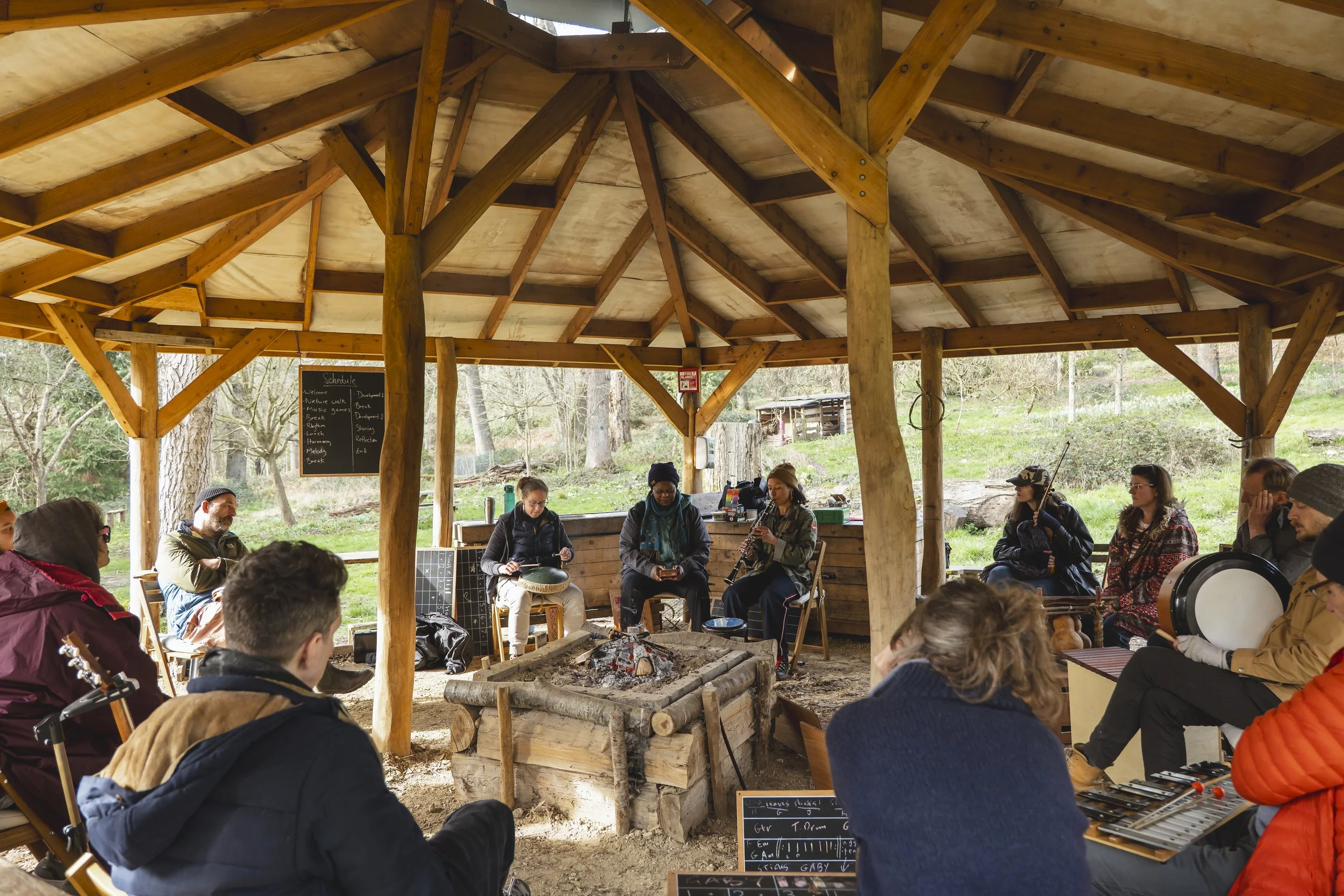 A group of people sitting around a campfire inside an outdoor shelter, with some playing musical instruments, during daytime.