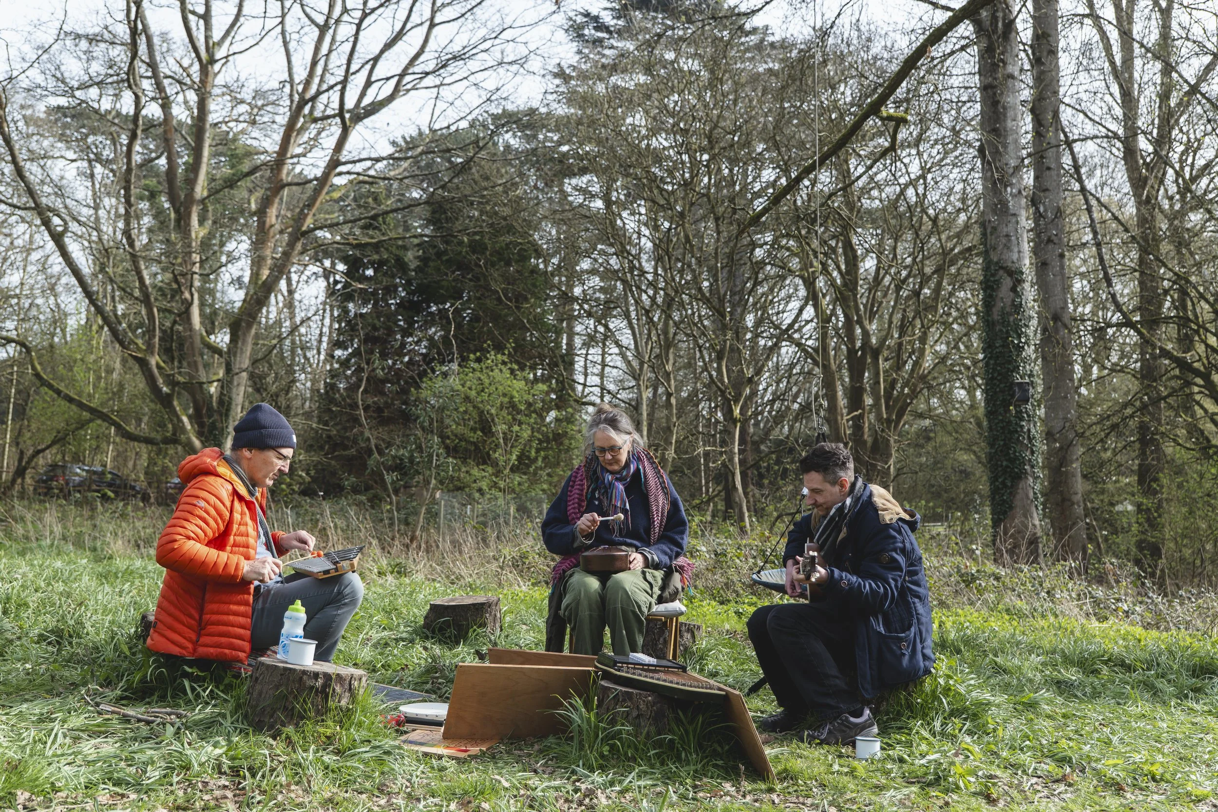 Three people sitting outdoors in a wooded area, playing musical instruments and singing, with various equipment and a box on the ground.