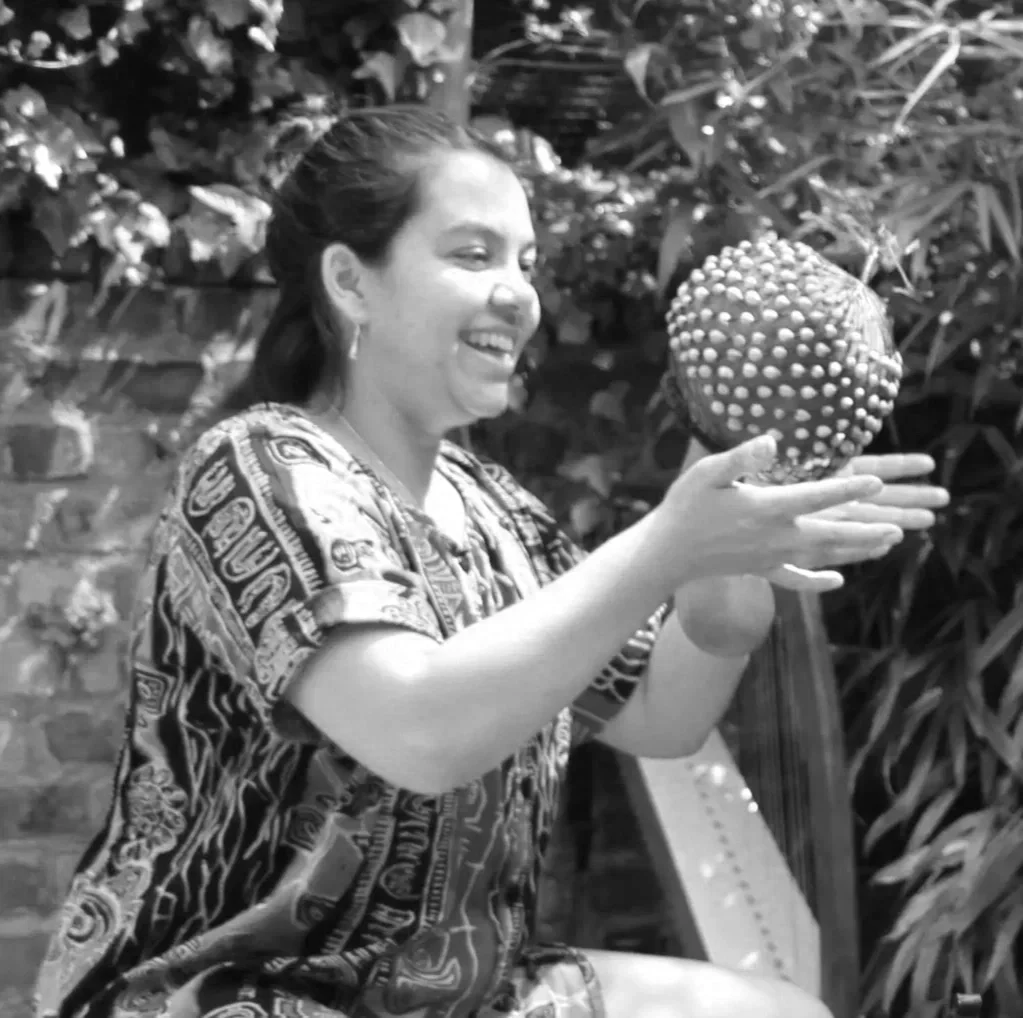 A woman smiling and holding a large, spiky fruit in her outstretched hand outdoors.