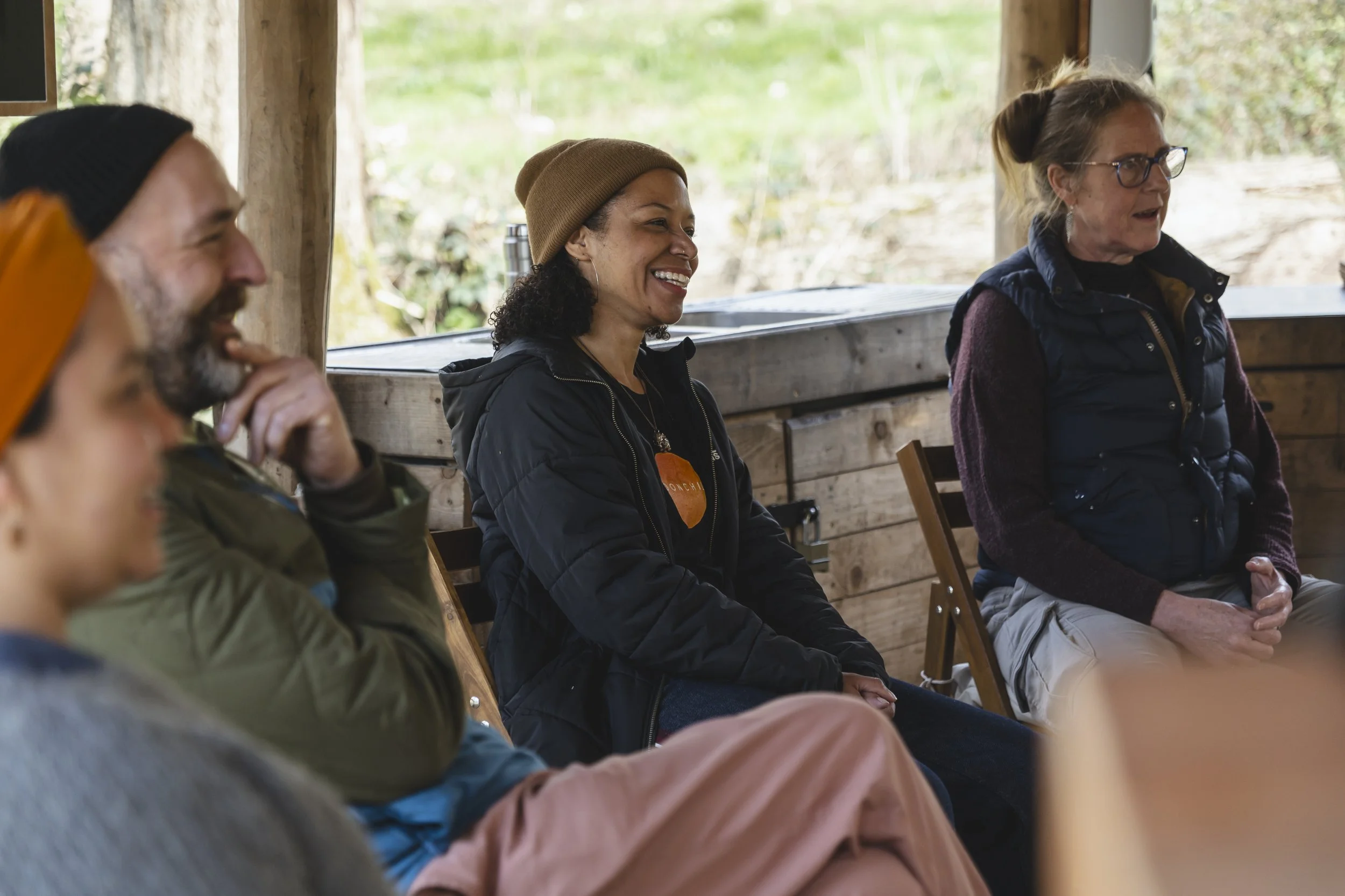 Group of smiling people sitting in wooden chairs indoors near a rustic wooden wall with greenery outside.