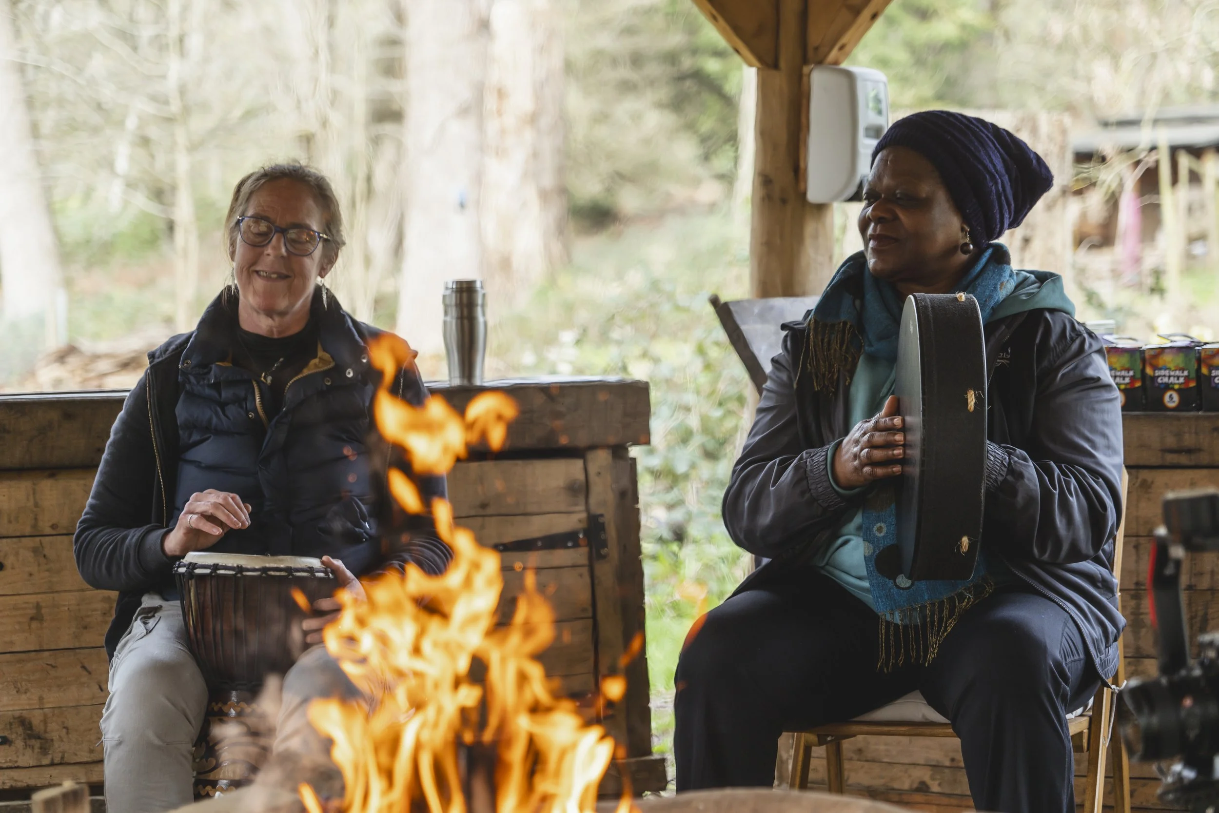 Two women sitting near a campfire, one playing a drum and the other holding a guitar case, in an outdoor shelter with trees in the background.
