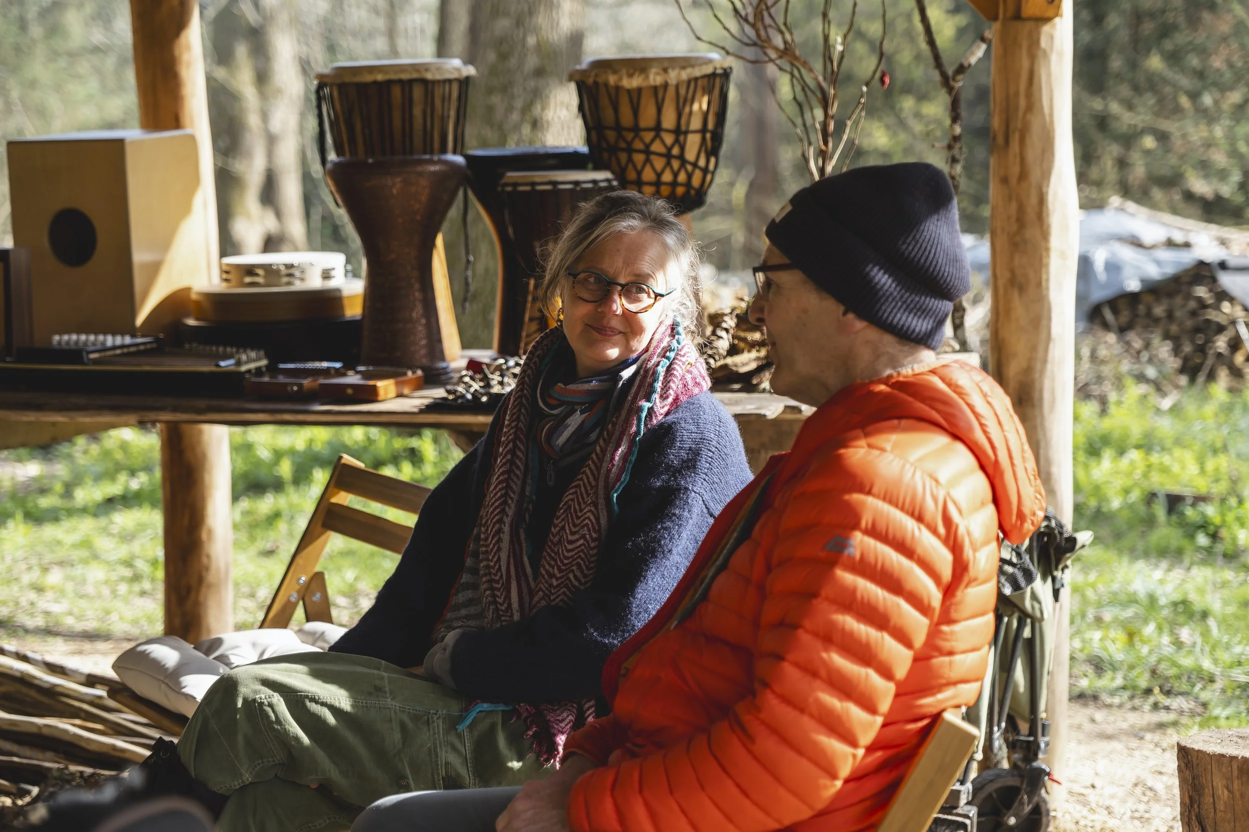 Two people sitting outdoors in a wooded area, conversing. A woman with blonde hair, glasses, dark sweater, and colorful scarf, and a man wearing a black beanie and an orange jacket. Behind them is a wooden table with African drums and other items.