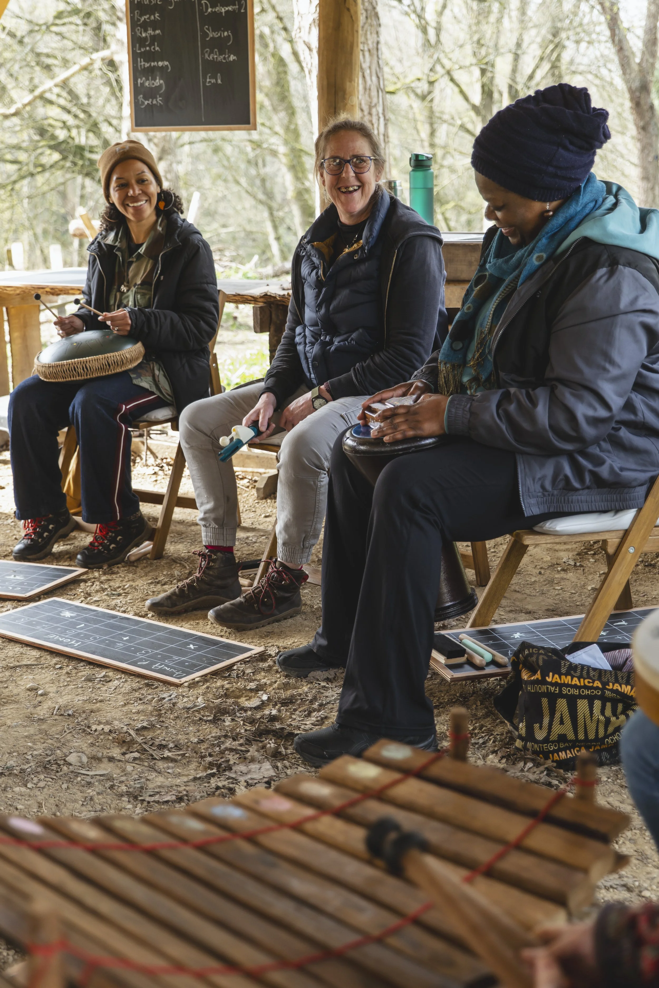 Three women sitting outdoors with musical instruments, smiling and engaging in a music activity, with chalkboards and educational materials on the ground and a wooden structure surrounding them.