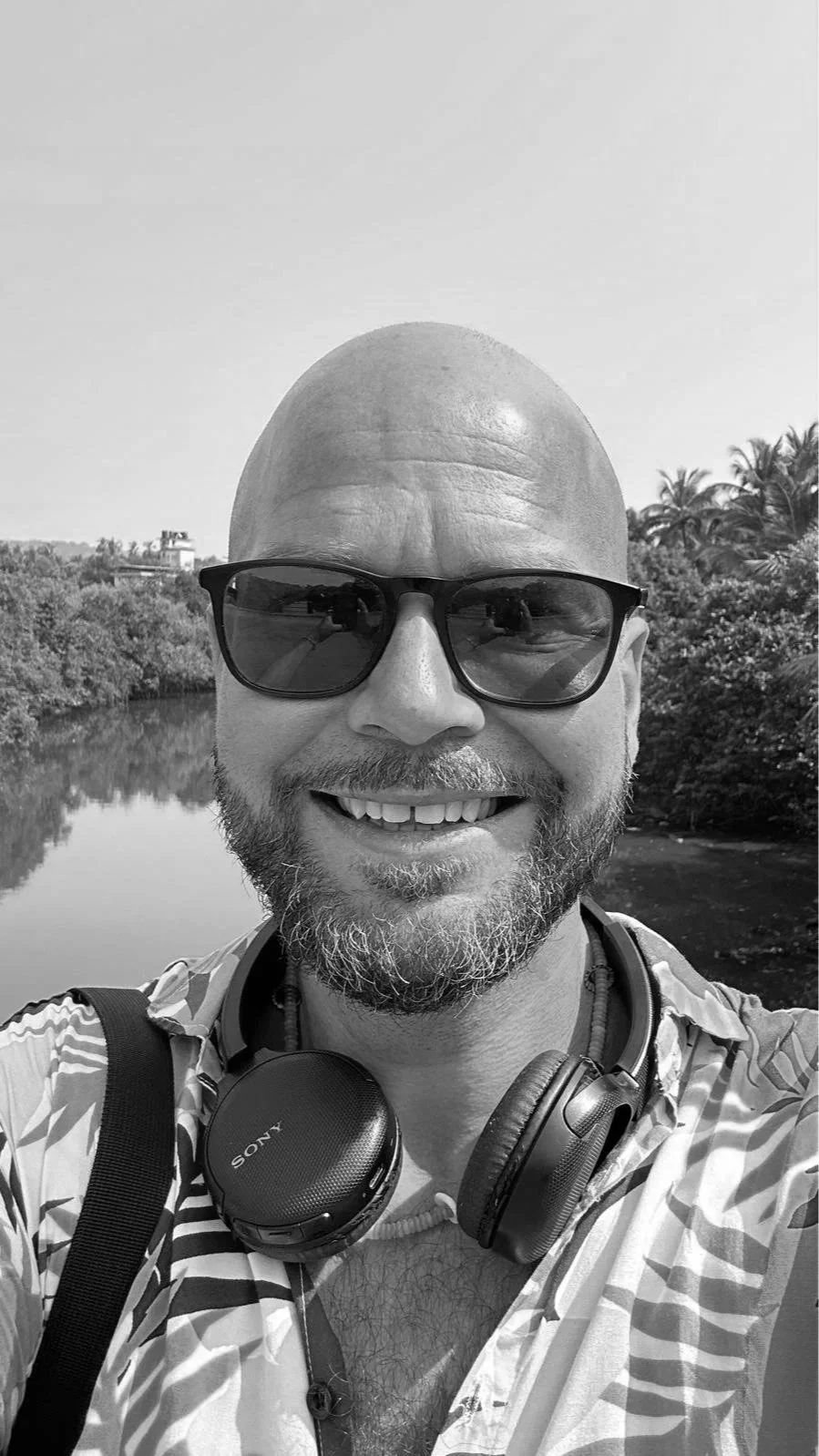 Black and white photo of a smiling man with a bald head, wearing sunglasses, headphones around his neck, and a striped shirt, outdoors near a body of water with trees and a building in the background.