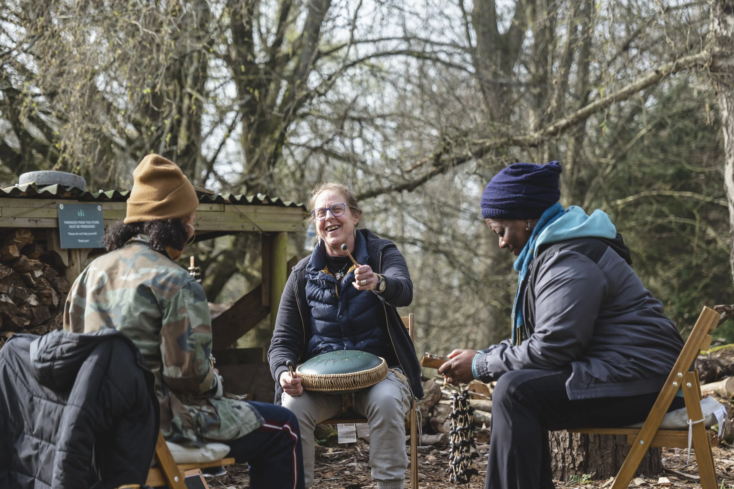 Three women sitting outdoors in a woodland area, engaging in music and conversation, with one woman holding a drum.