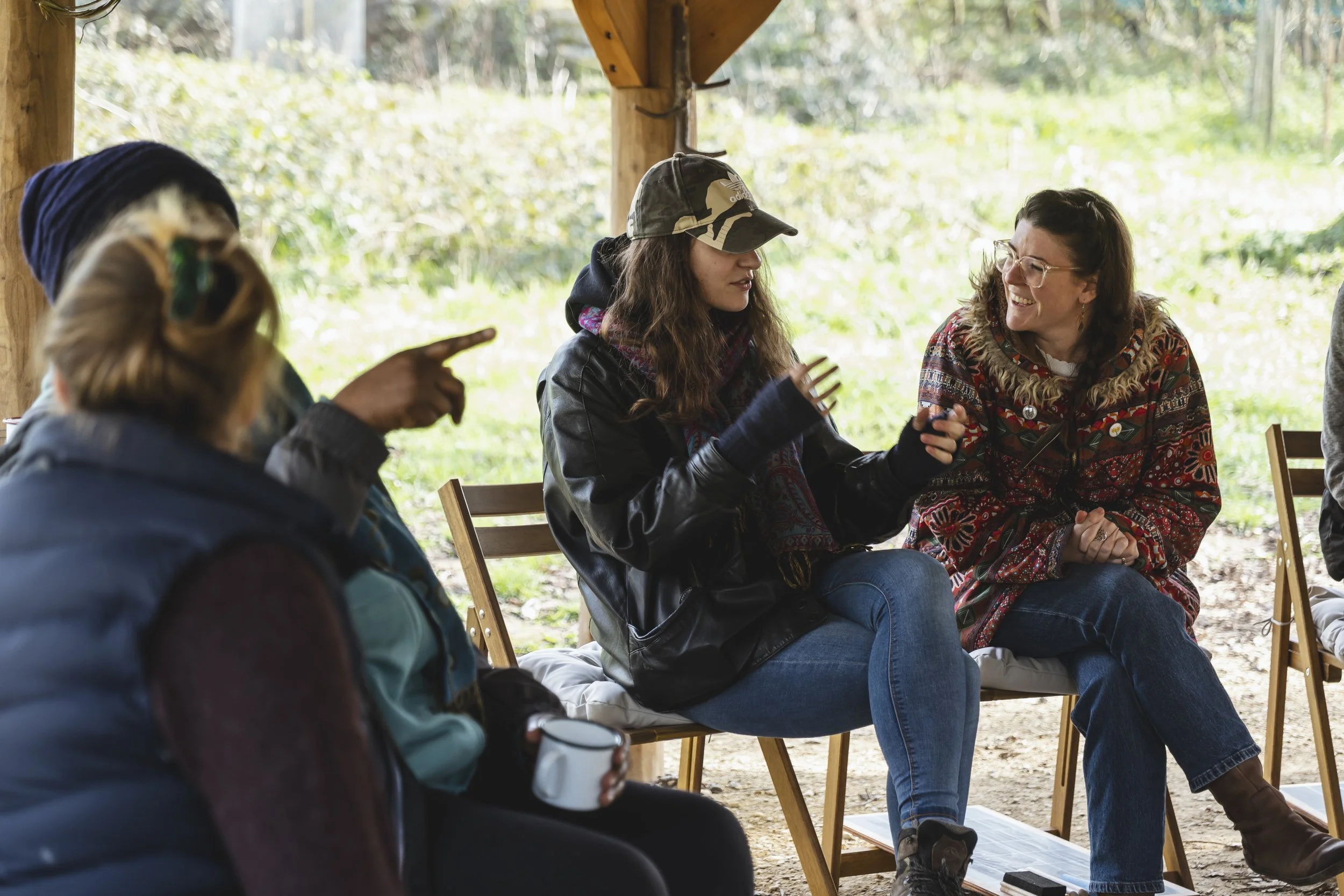 Group of people sitting on chairs under a wooden shelter, engaged in conversation, with greenery in the background.