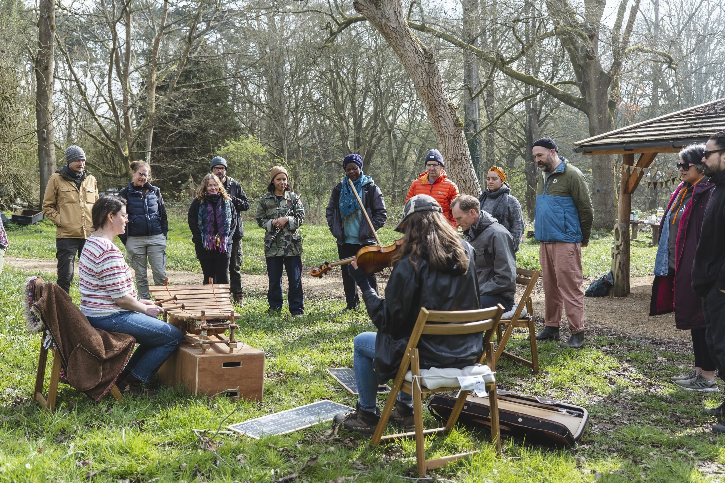 Group of people gathered outdoors in a park-like setting, some watching two musicians playing instruments, a violin and a percussion instrument.