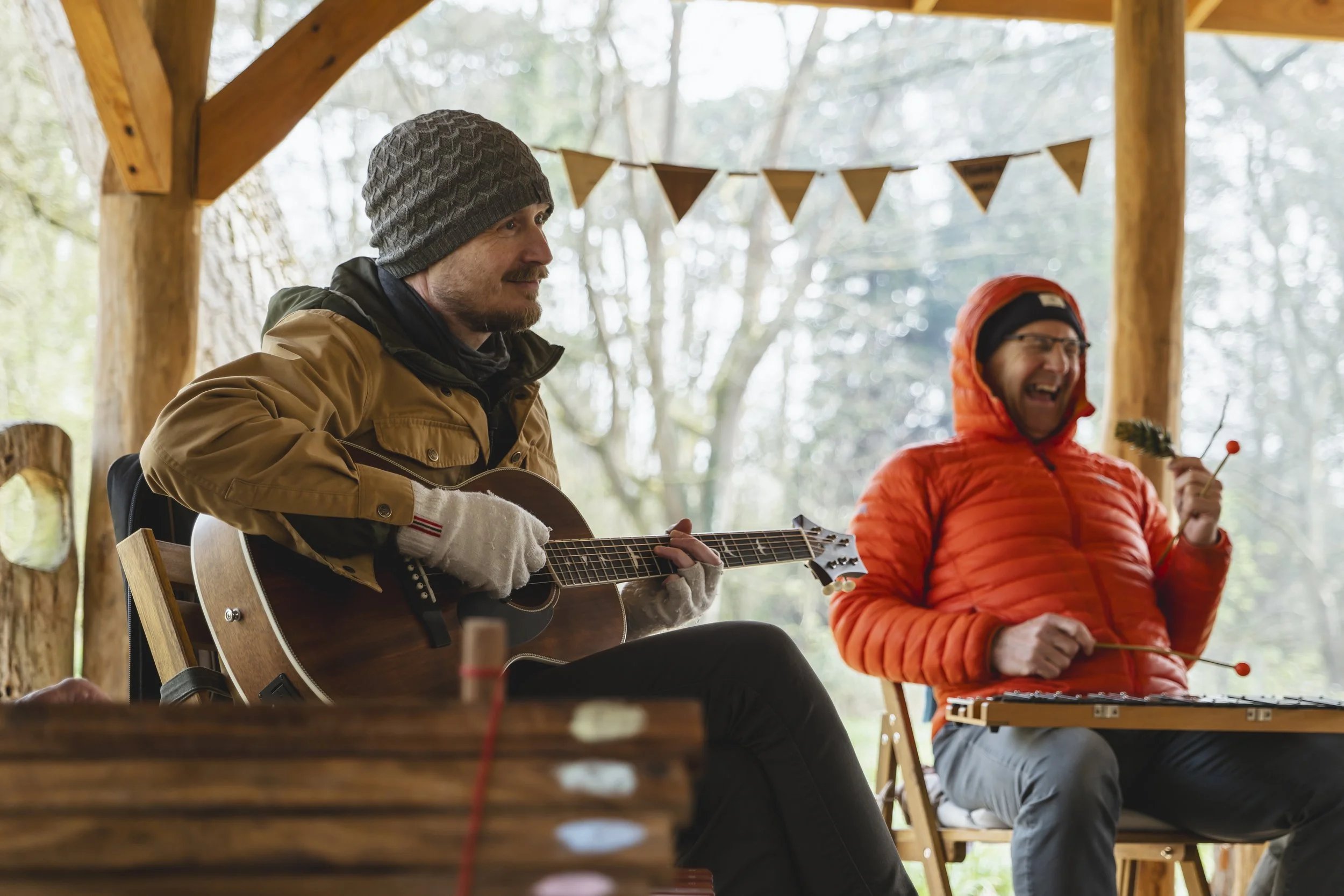 Two men sitting outdoors under a wooden shelter, playing guitar and xylophone, with triangular bunting hanging above them, surrounded by trees.