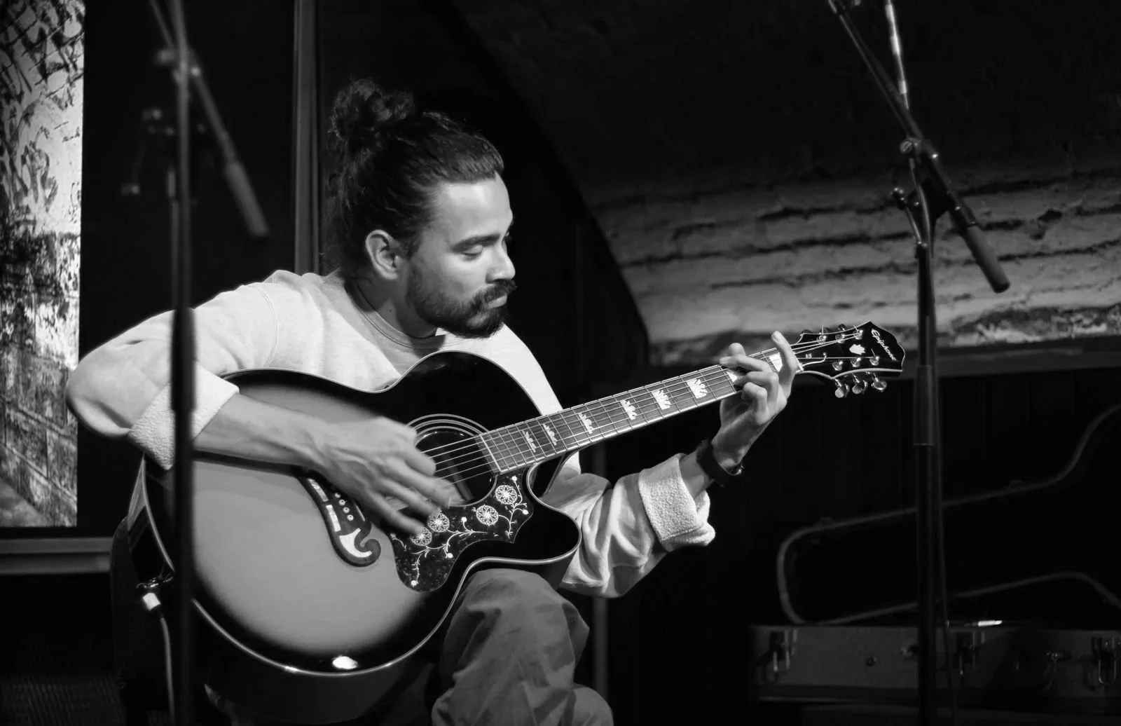 A man with long hair tied in a bun and a beard, playing an acoustic guitar on stage in a black and white photo.