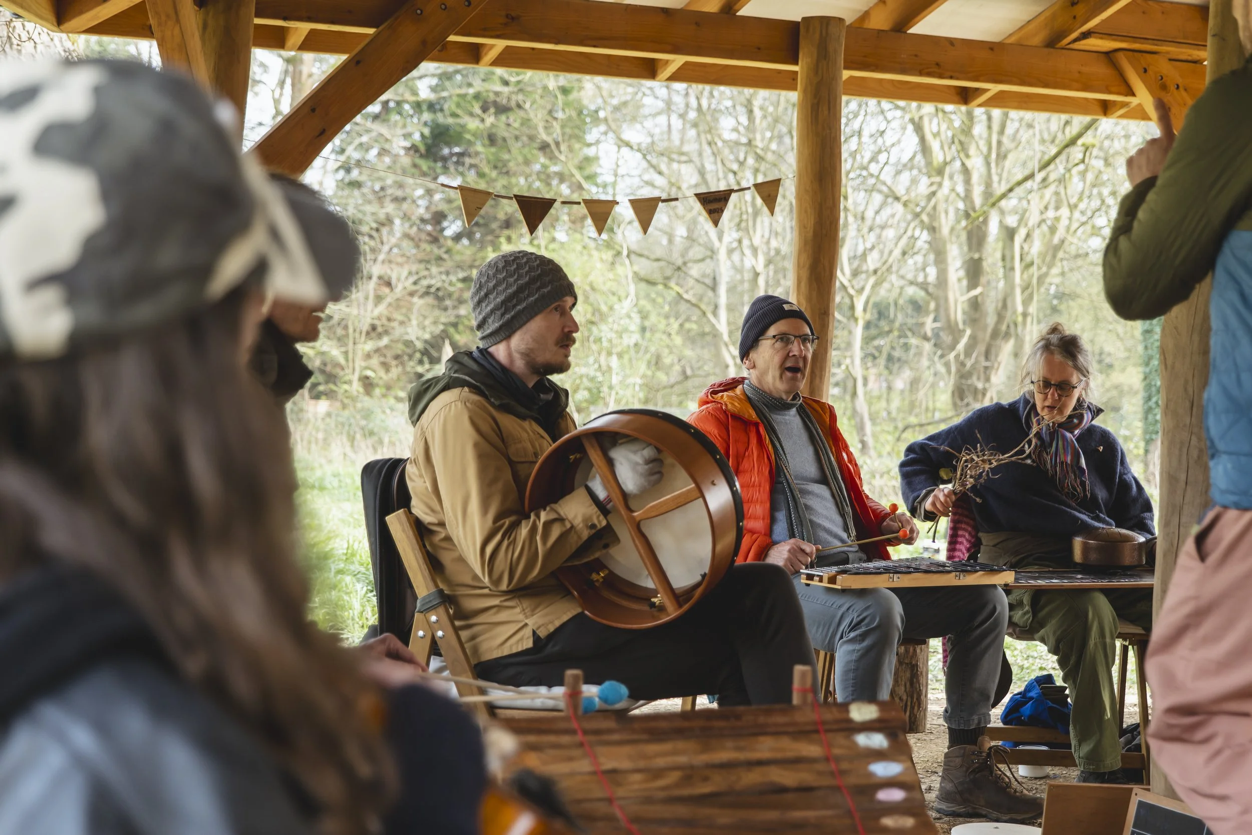 A group of people sitting under a wooden shelter in a forest, some playing musical instruments, including a drum and a flute, while others watch and listen.