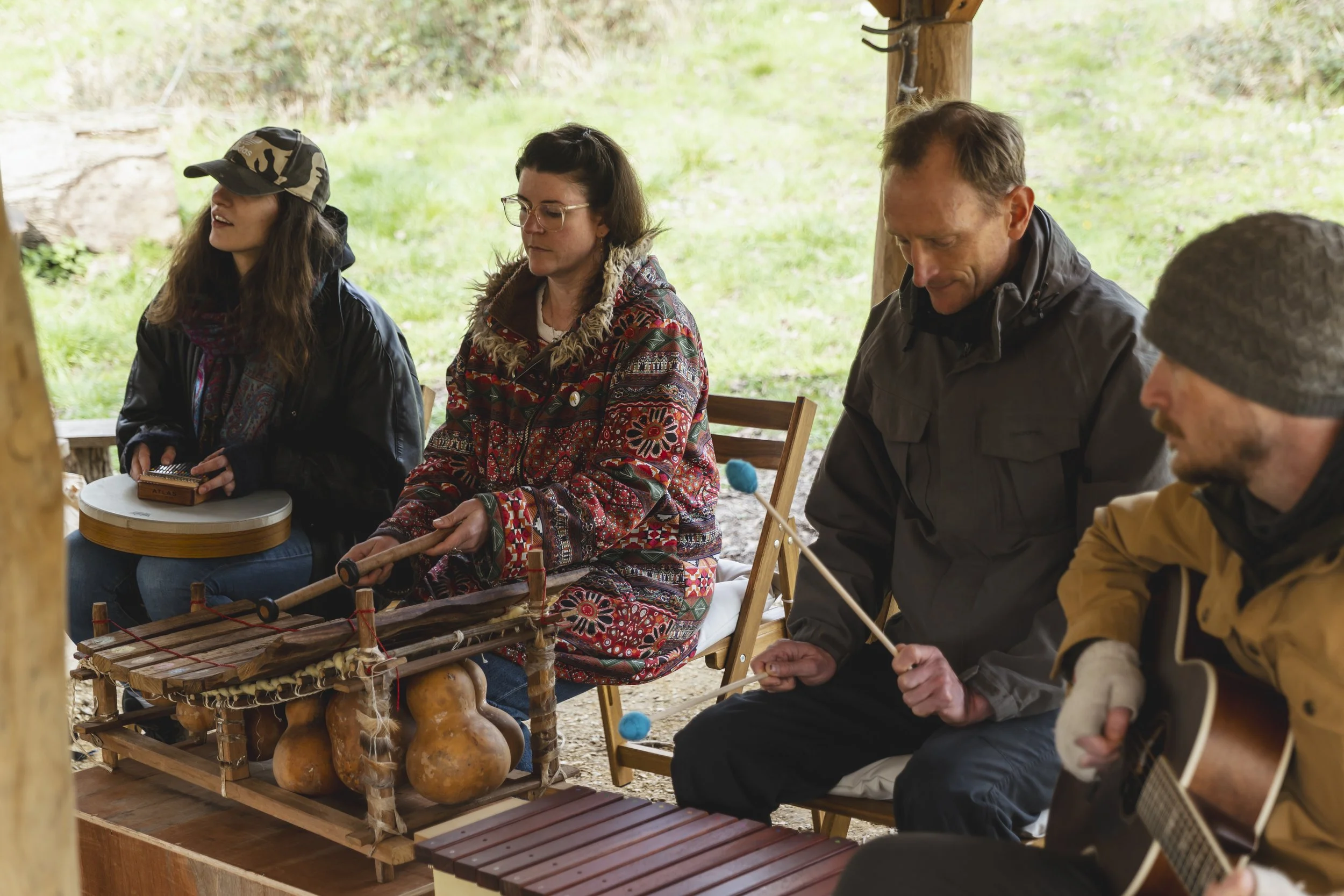 Four people sitting outdoors, playing musical instruments and holding percussion mallets, under a wooden shelter with a grassy background.