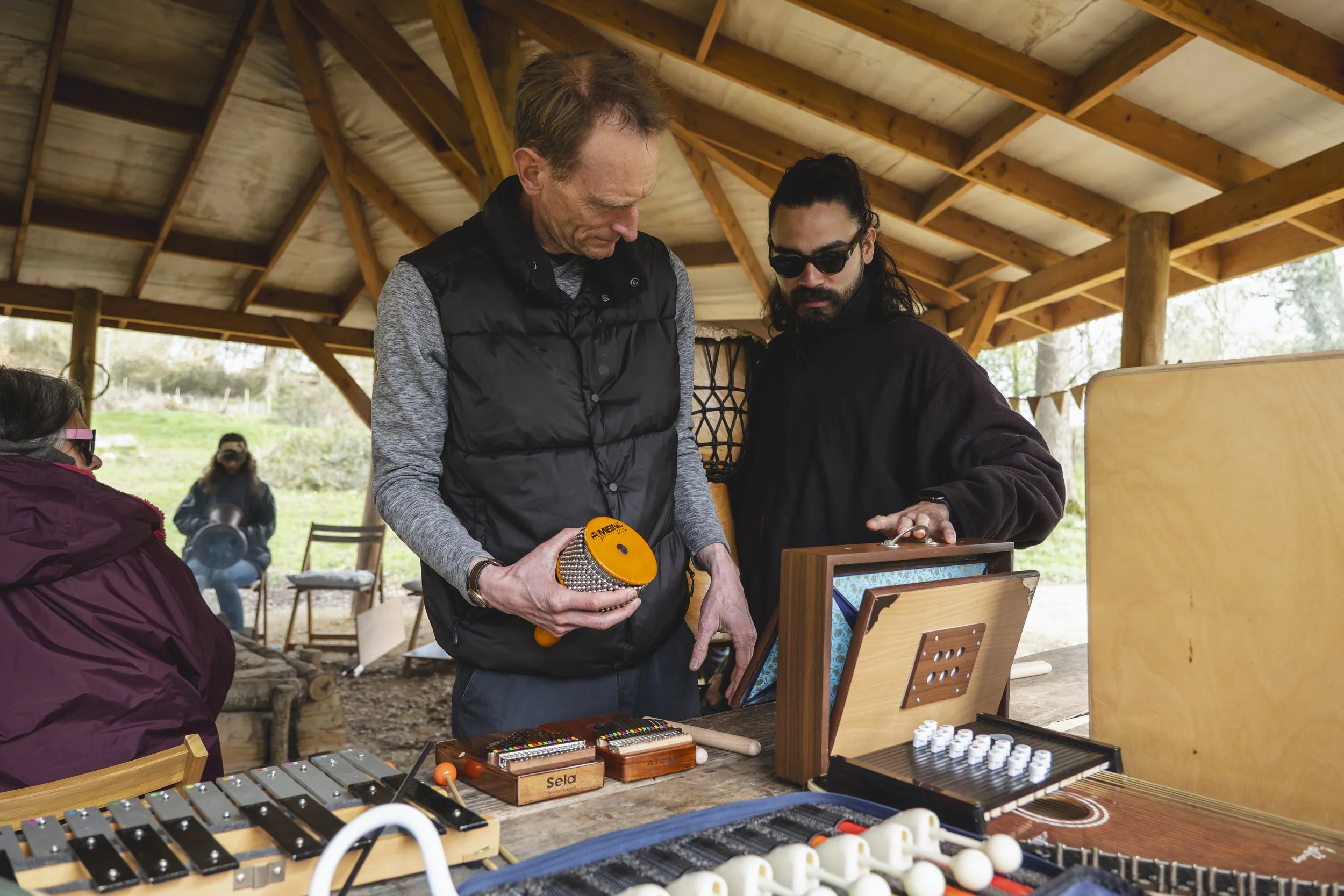 Two men are examining musical instruments on a wooden table under a wooden roofed outdoor setting. The man on the left holds a small orange percussion instrument, while the man on the right wears sunglasses and looks at the instrument.