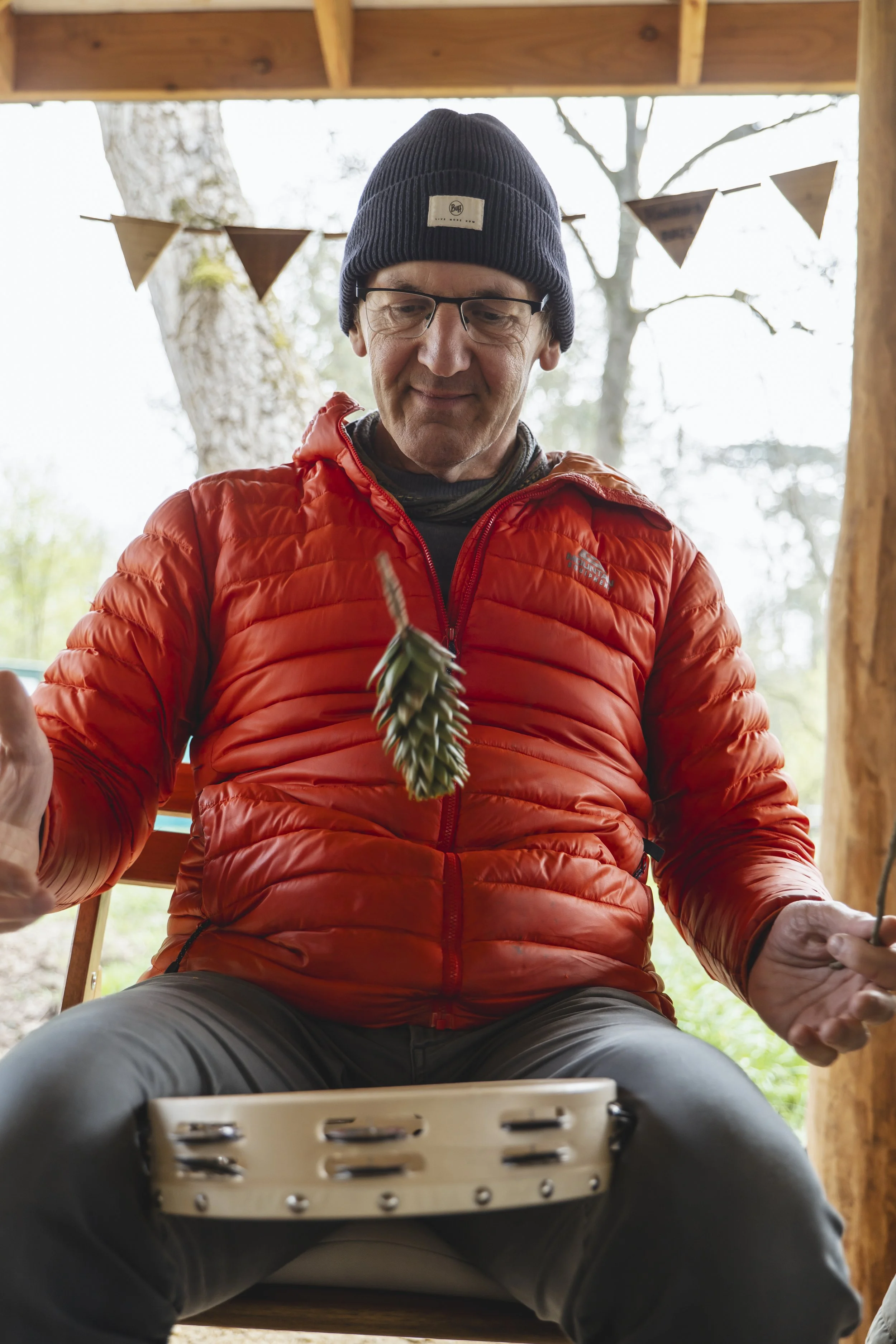 A man sitting outdoors under a wooden shelter, wearing a black beanie, glasses, an orange puffer jacket, and gray pants. He is playing a tambourine with pine cones attached and has a small pine cone hanging from his neck.