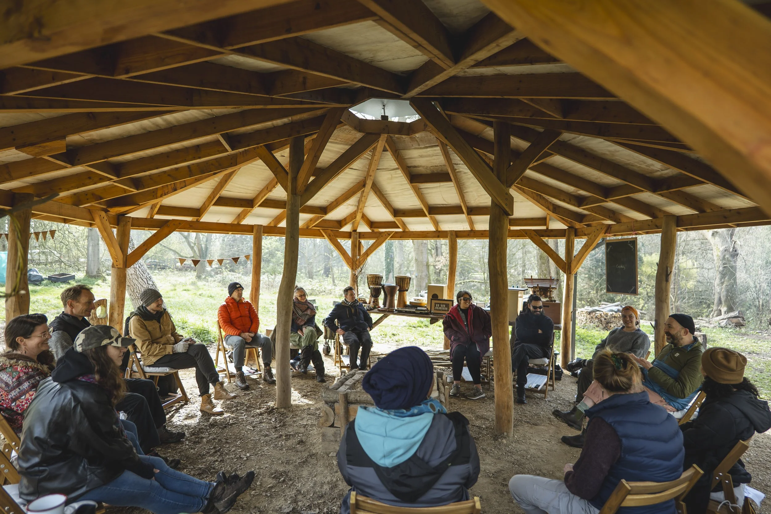 A group of people seated in a circle inside a wooden, open-air pavilion surrounded by trees, participating in a discussion or workshop.