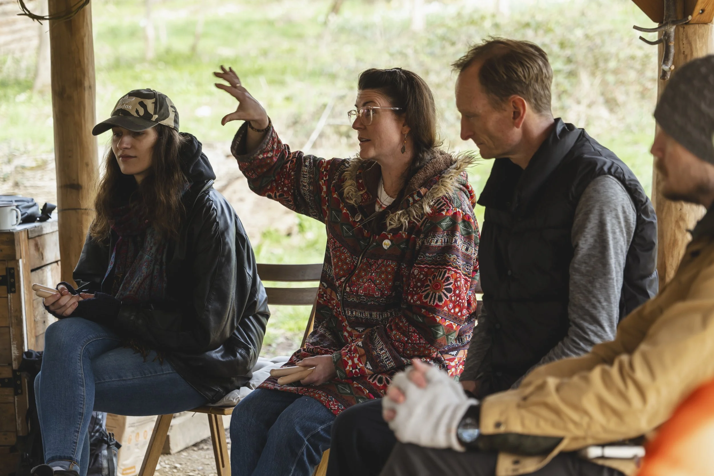 Group of people sitting and listening to a woman speaking, outdoors in a shelter with wooden posts and greenery in the background.