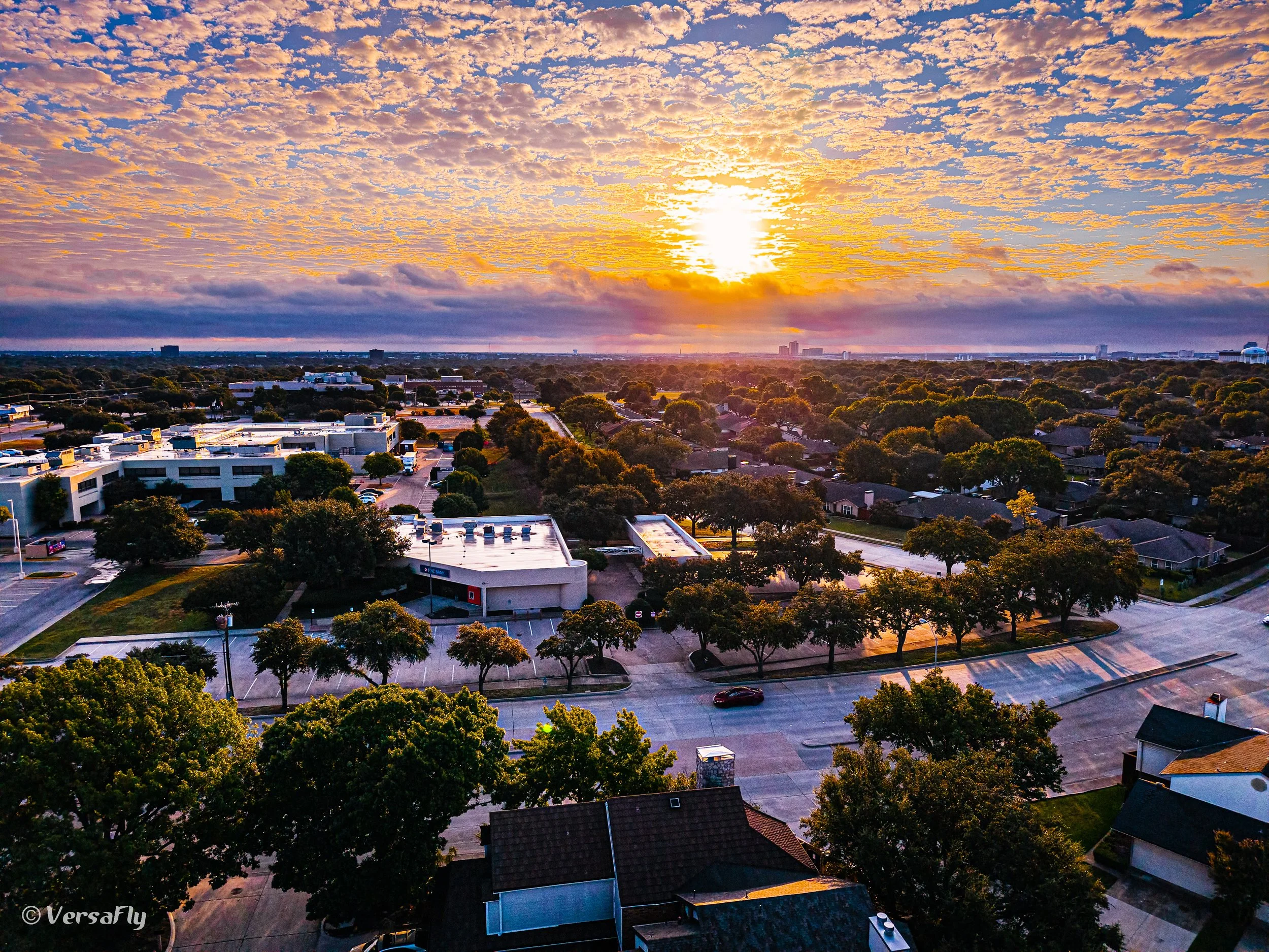 A cityscape at sunset with a sky filled with orange and pink clouds, residential houses, a parking lot, and a commercial building with a parking lot in the foreground, and trees lining the streets.