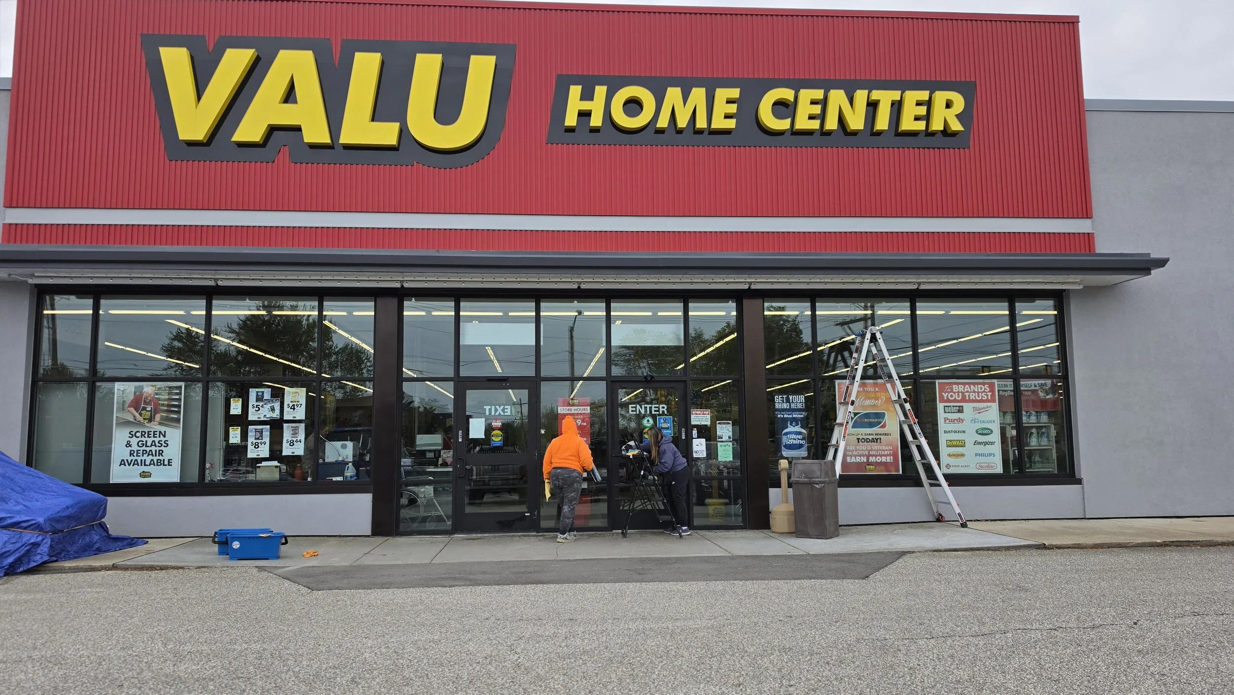 Front of a Valu Home Center store with large yellow sign. Two people are standing outside the entrance, one in an orange hoodie and the other in a purple jacket, with ladder and boxes nearby. Store windows display various advertisements and signs.