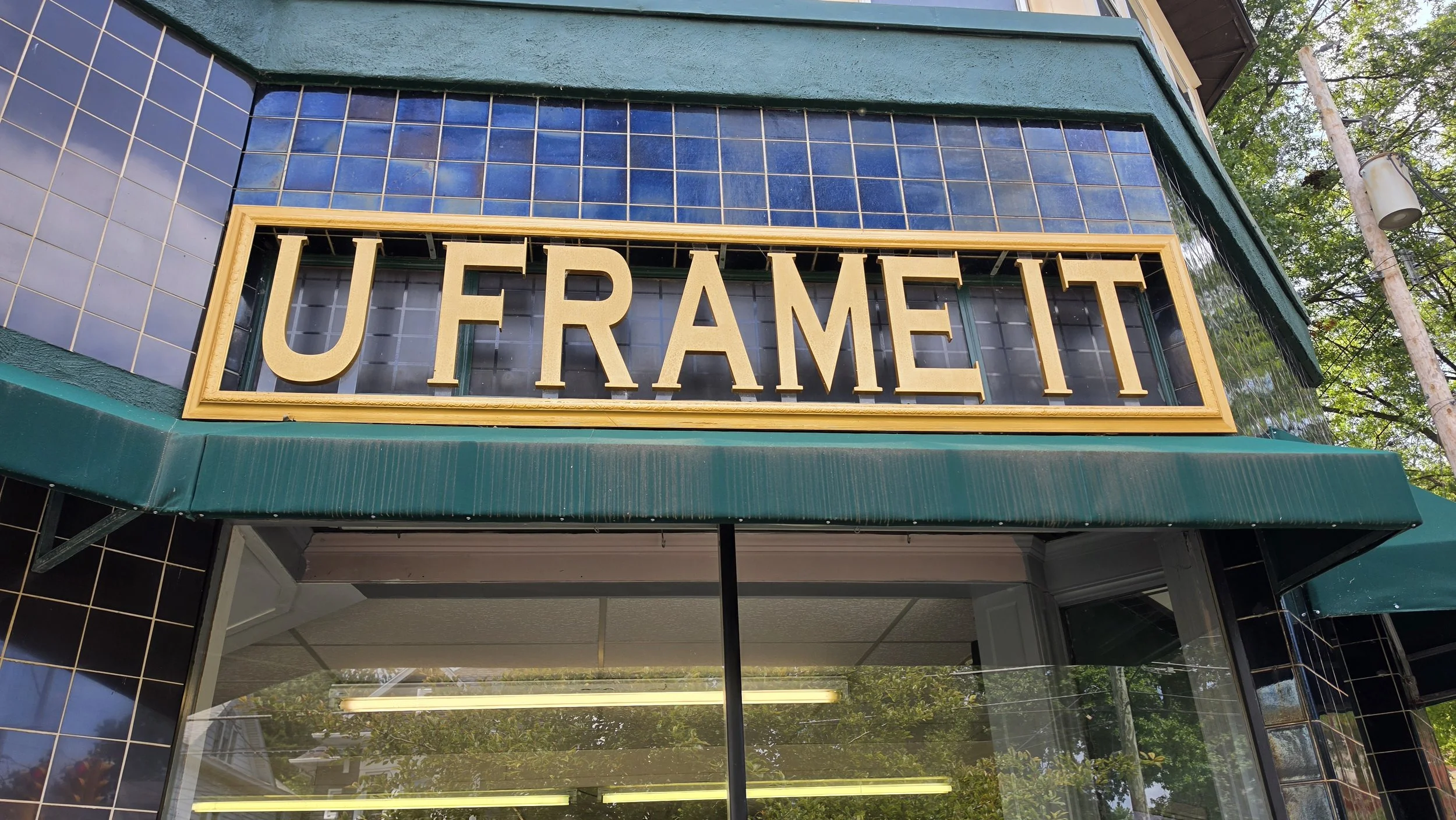 Close-up of a store sign that reads 'UFRAME IT' in large, gold letters, attached to a building with black tile exterior and a green awning.