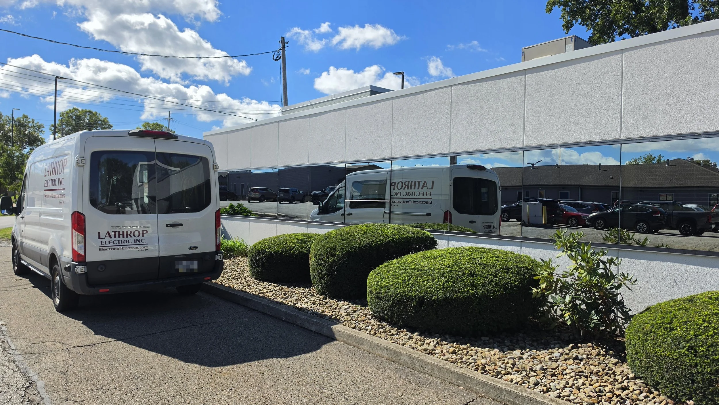 A white van with the logo 'Lathrop Electric Inc.' parked near a white building with reflective windows. The background features a blue sky with clouds, trees, power lines, and parked cars in a parking lot.