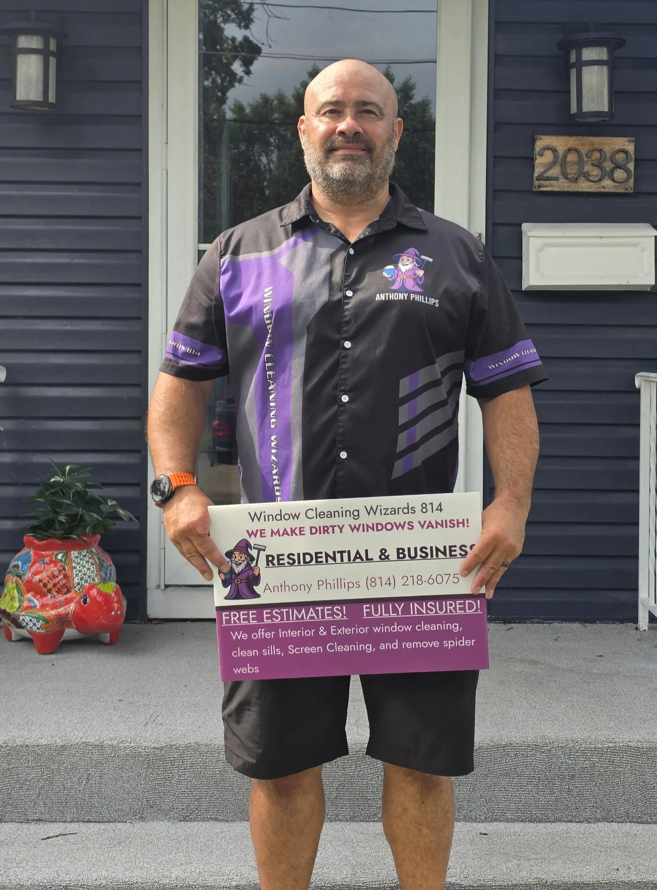 A man standing in front of a house holding a sign advertising window cleaning services. The man is wearing a black shirt with purple accents and the name Anthony Phillips. The house has the address 2038 on a plaque, and there is a decorative piggy bank on the porch near a potted plant.