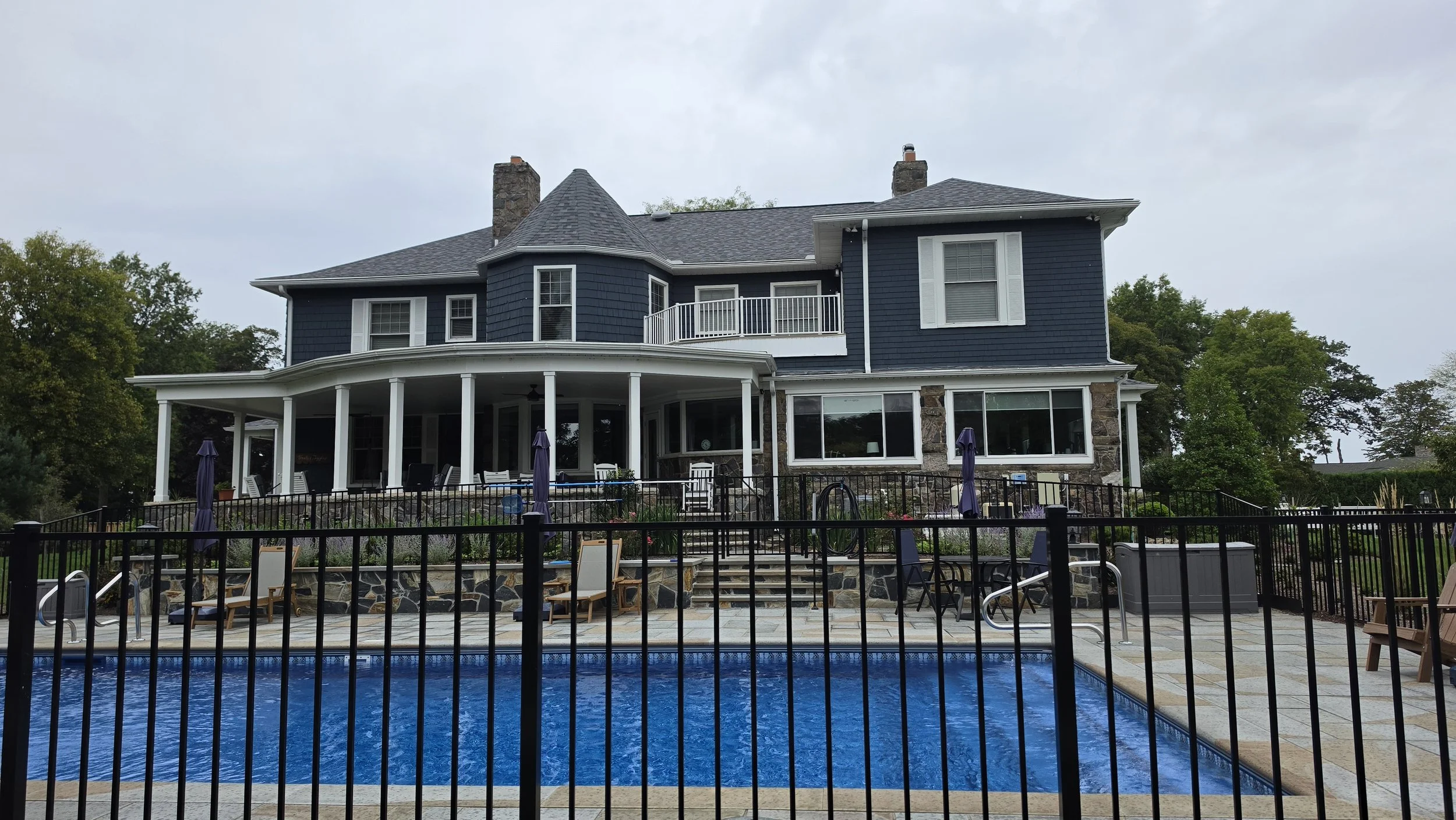 A large blue house with multiple windows, a balcony, and a covered porch, surrounded by trees. In the foreground, there is a swimming pool with a black fence, poolside chairs, umbrellas, and outdoor furniture.