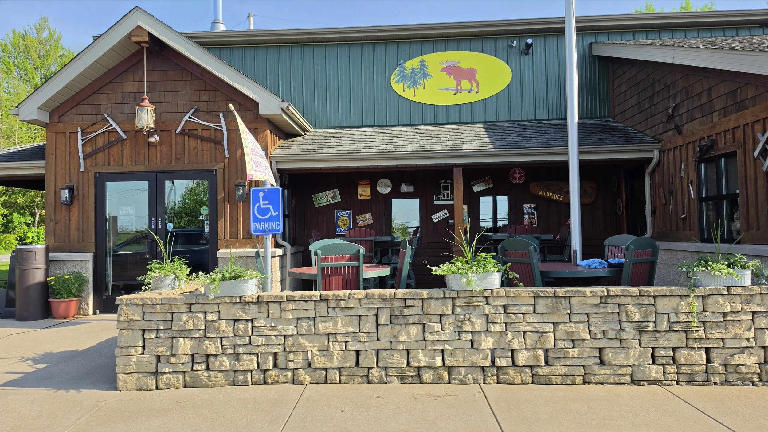 Exterior of a rustic restaurant or café with outdoor seating, potted flowers, a stone wall, and decorative outdoor elements, including an eagle and moose logo sign on the building.