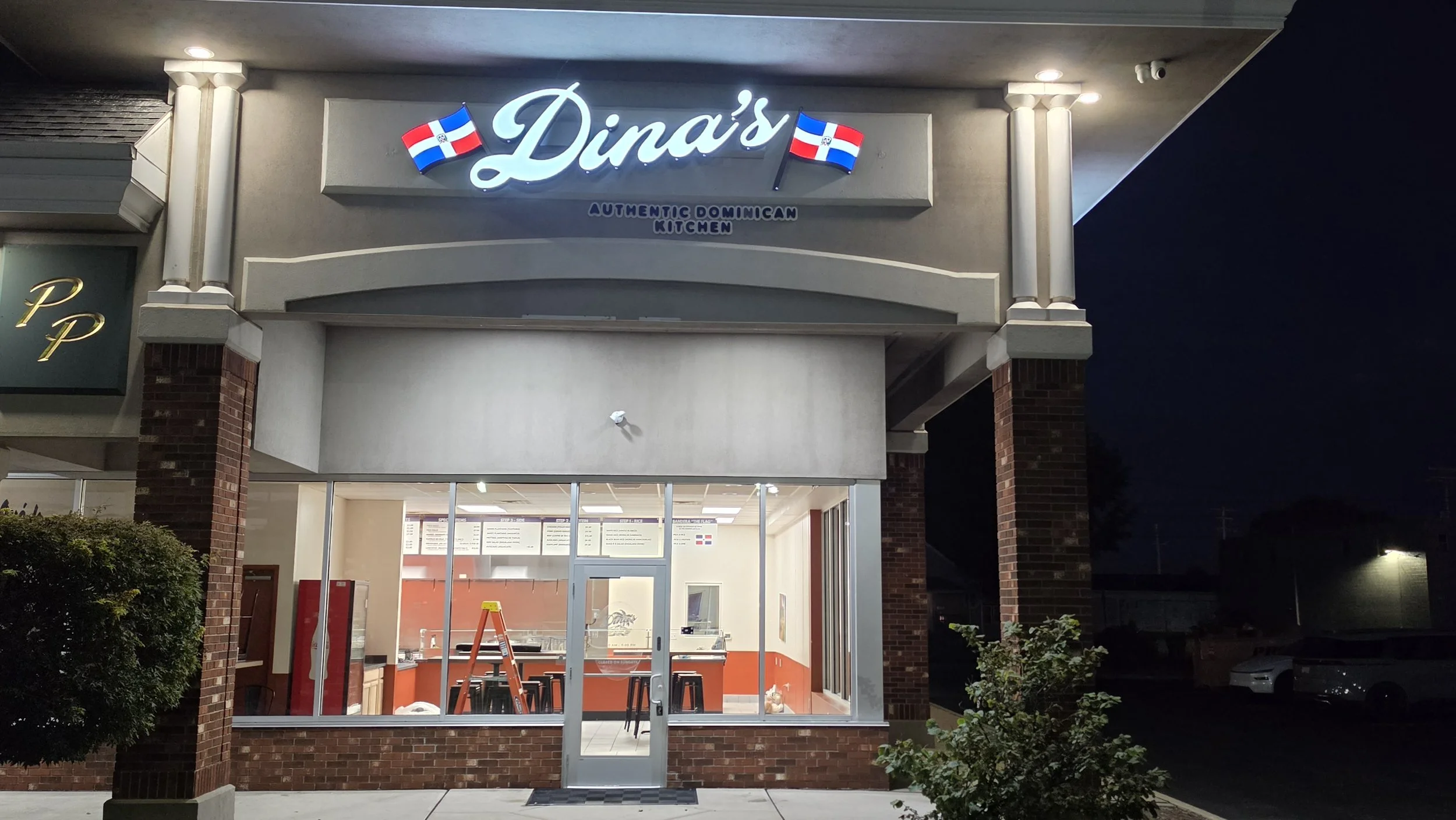 Front of a restaurant named Dina's with illuminated sign and Dominican flags, photo taken at night. The restaurant has a large glass window and a door, with a small menu visible inside.