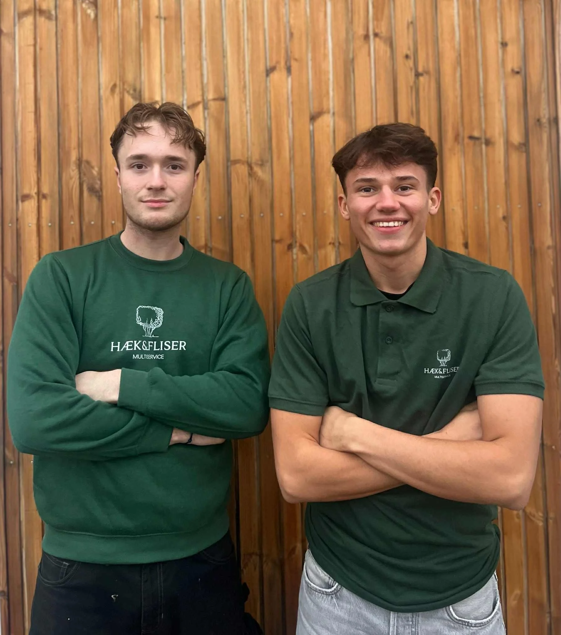 Two young men standing with arms crossed in front of a wooden wall, wearing green shirts with a tree logo and the text 'HÆK & FLISER MULTISERVICE'.