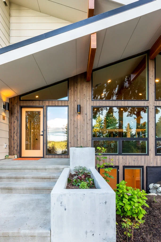 Modern house entrance with concrete stairs, wooden exterior walls, and large glass windows reflecting trees. There is a white planter with plants at the base of the stairs and some green shrubbery nearby.