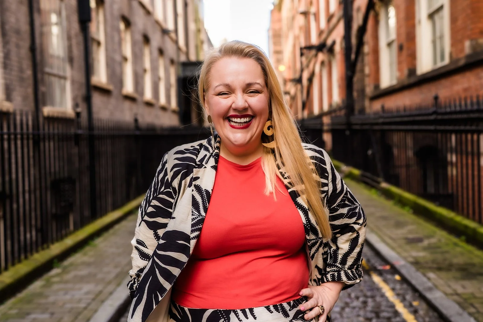 Lois Bates-Stubbs, a blond woman in her early forties is smiling at the camera, her left hand on her hip. She is wearing a leaf pattern suit in black & cream with a ref t-shirt, red lipstick and big gold earrings. She is stood on a cobbled street.