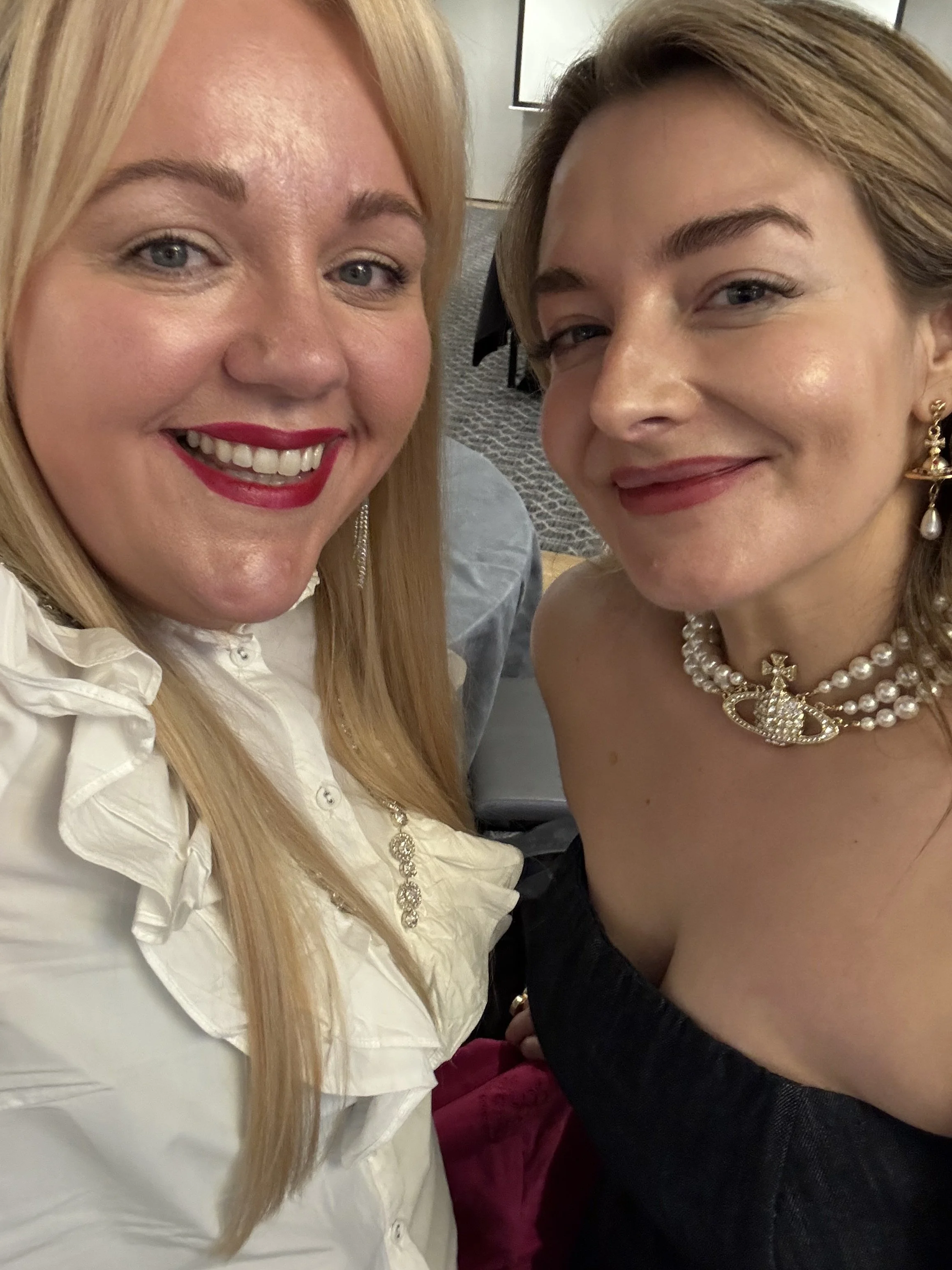 Two women, one blonde, one brunette are smiling side by side. It's a selfie. They both look happy and are well dressed, at a business event.