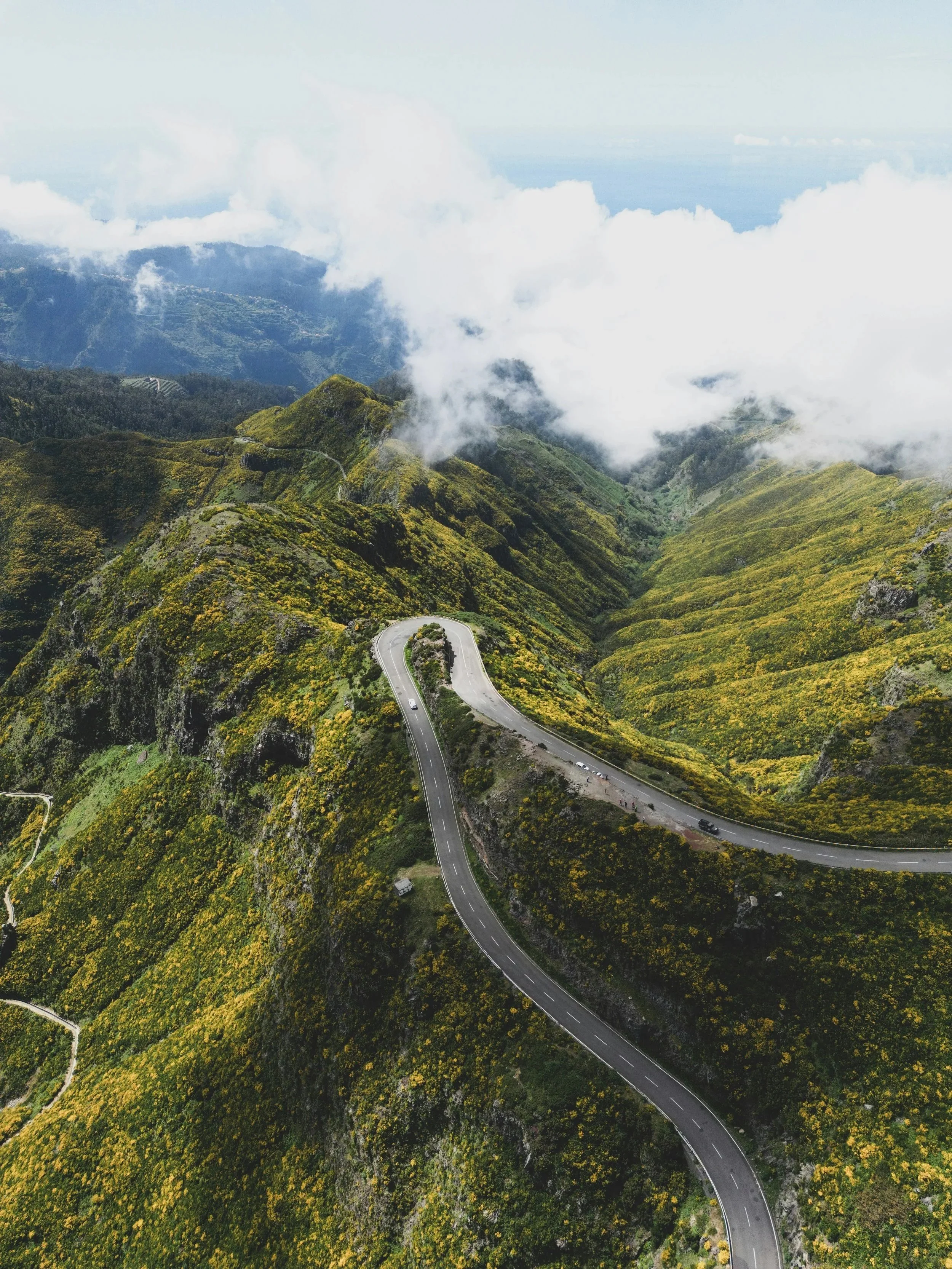 A winding mountain road surrounded by green hills and forests, with clouds covering the mountain peaks,  Madeira Island, Madeira in style.