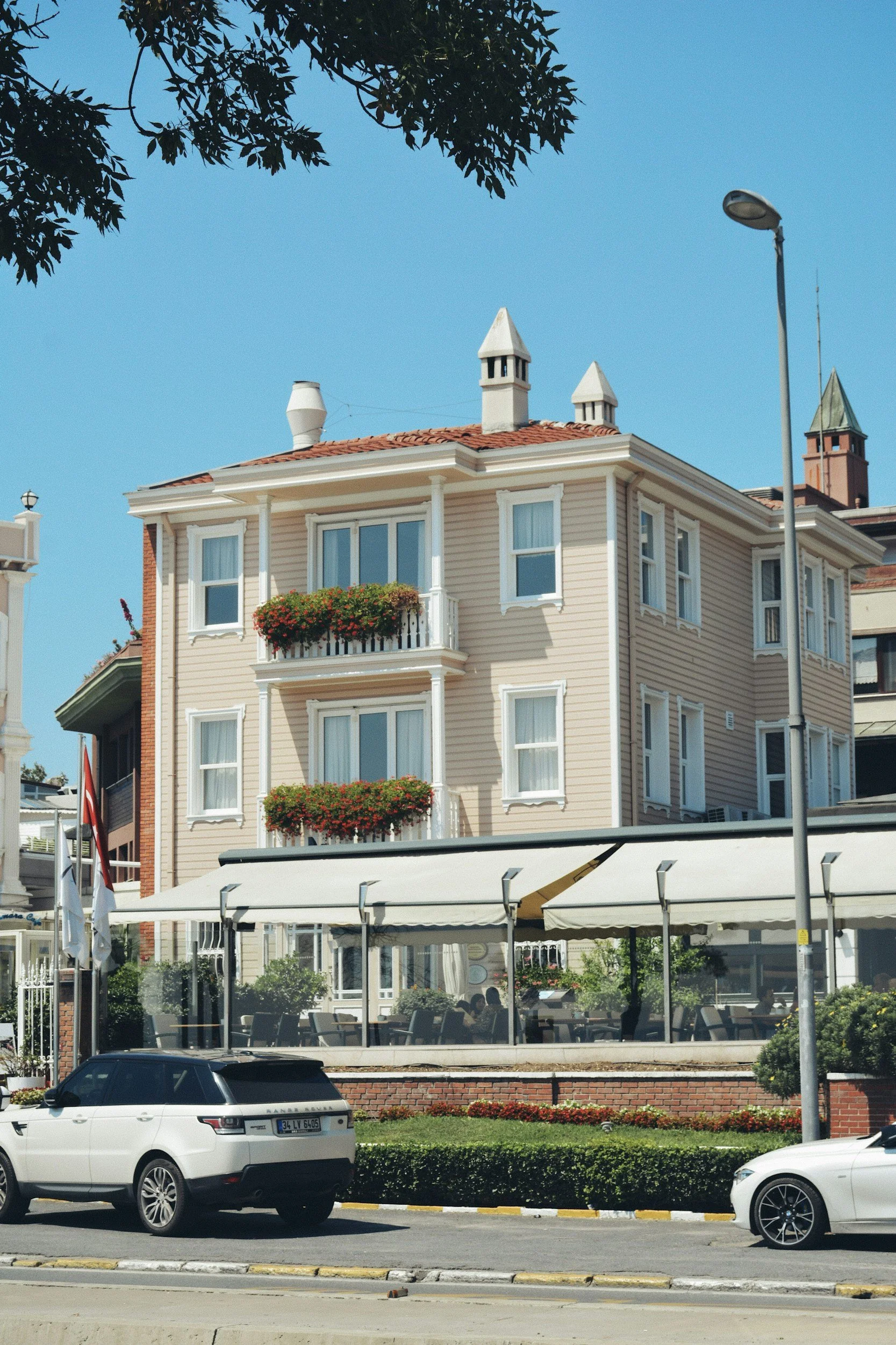 A multi-story building with cream-colored siding, white trim, and red-tiled roof, featuring balconies with flower boxes, and a restaurant patio with outdoor seating in front, with parked cars along the street.