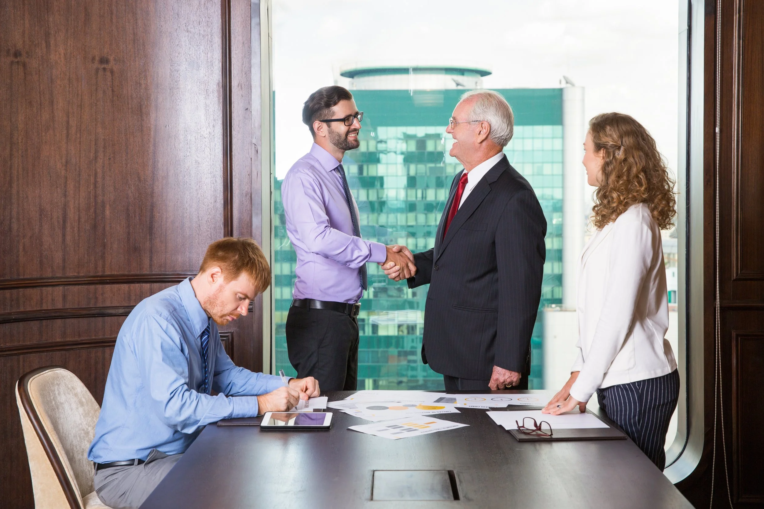 Two men in business suits shaking hands in an office with two women and a table with documents and electronic devices.