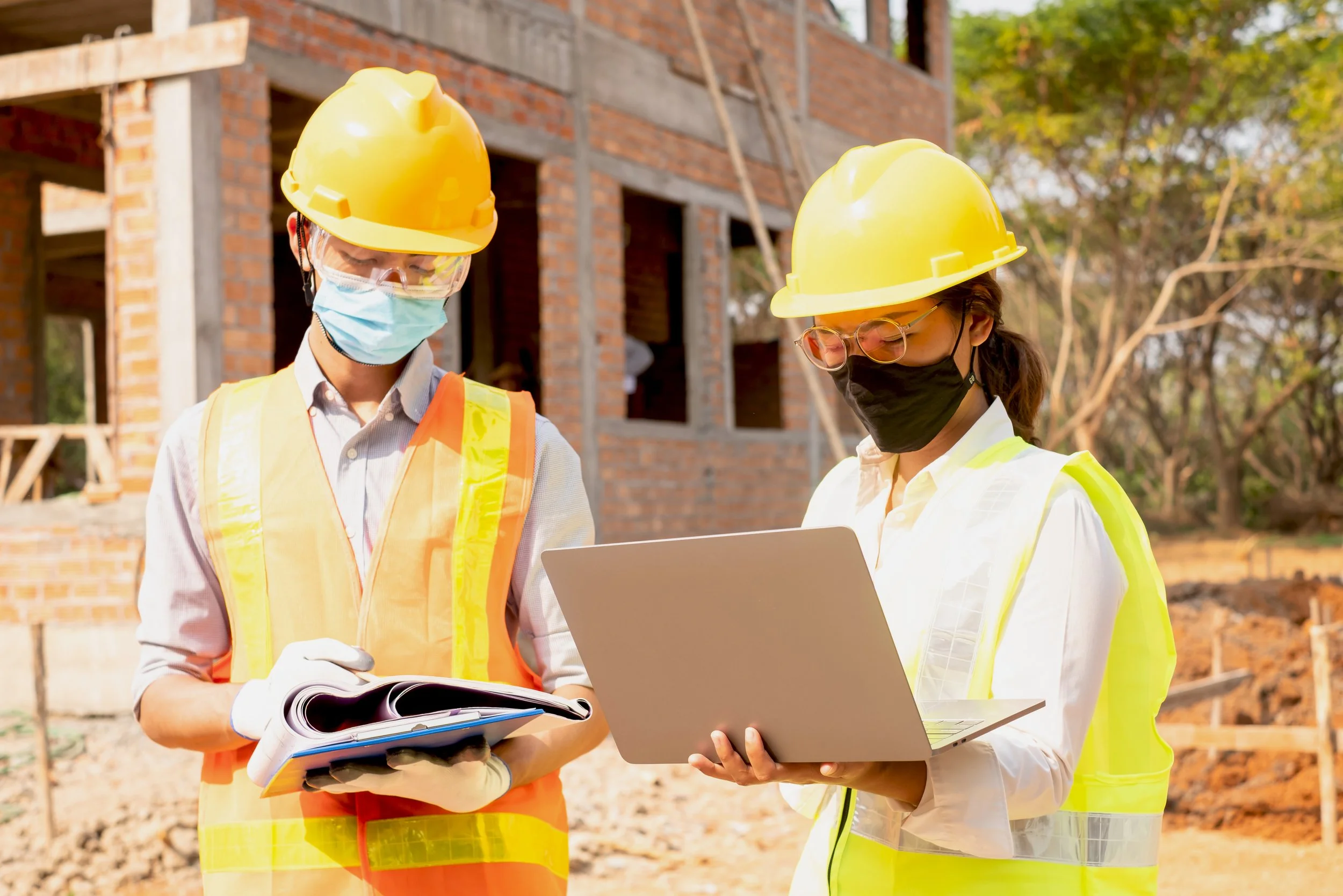 Two construction workers, a man and a woman, wearing yellow safety helmets, high-visibility vests, protective masks, and glasses, reviewing documents and a laptop at a building site with a brick building under construction in the background and trees.