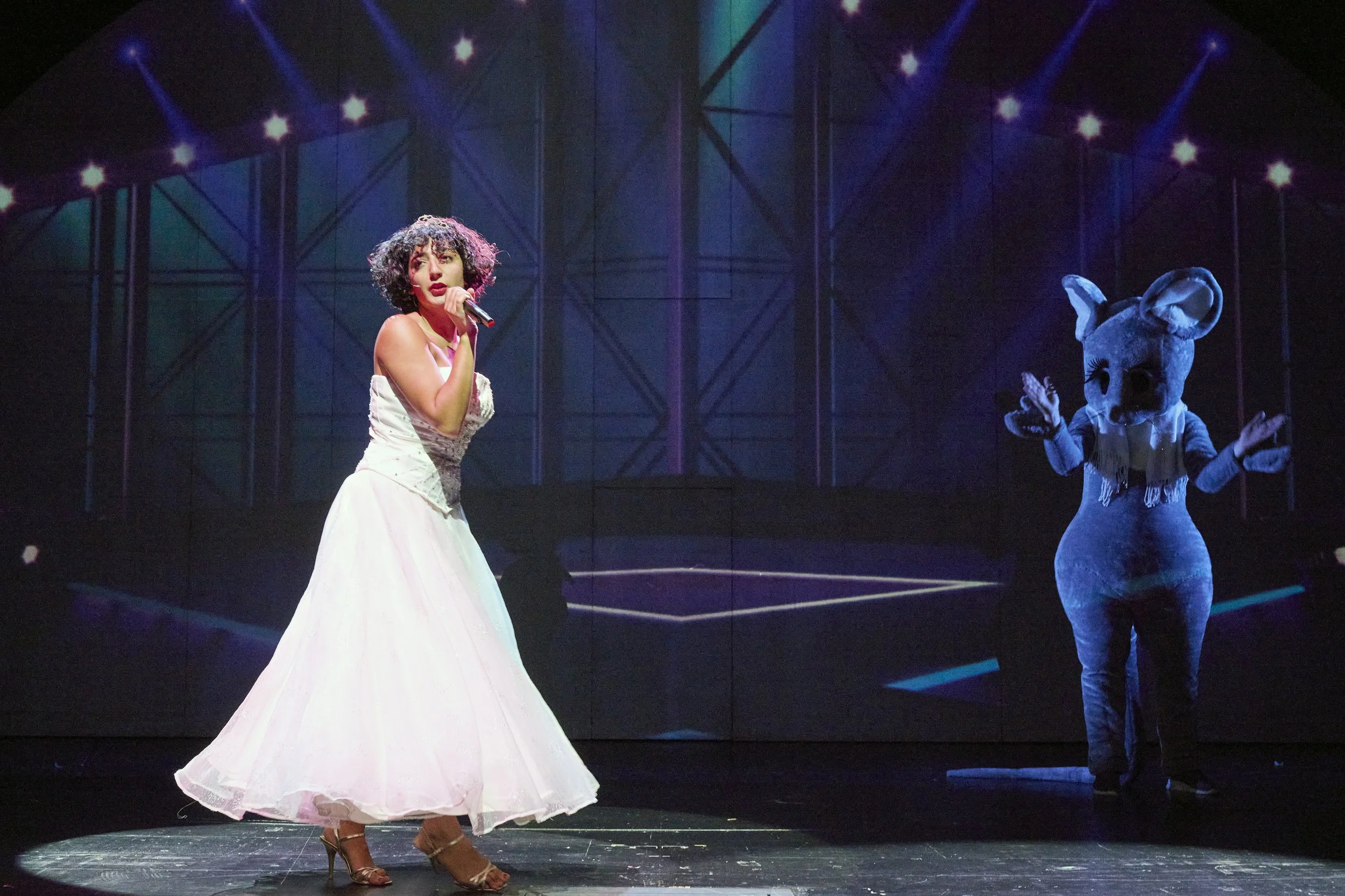 Eine Frau in einem langen weißen Kleid singt auf einer Bühne, neben einer Person in einem Tierkostüm mit großen Ohren.