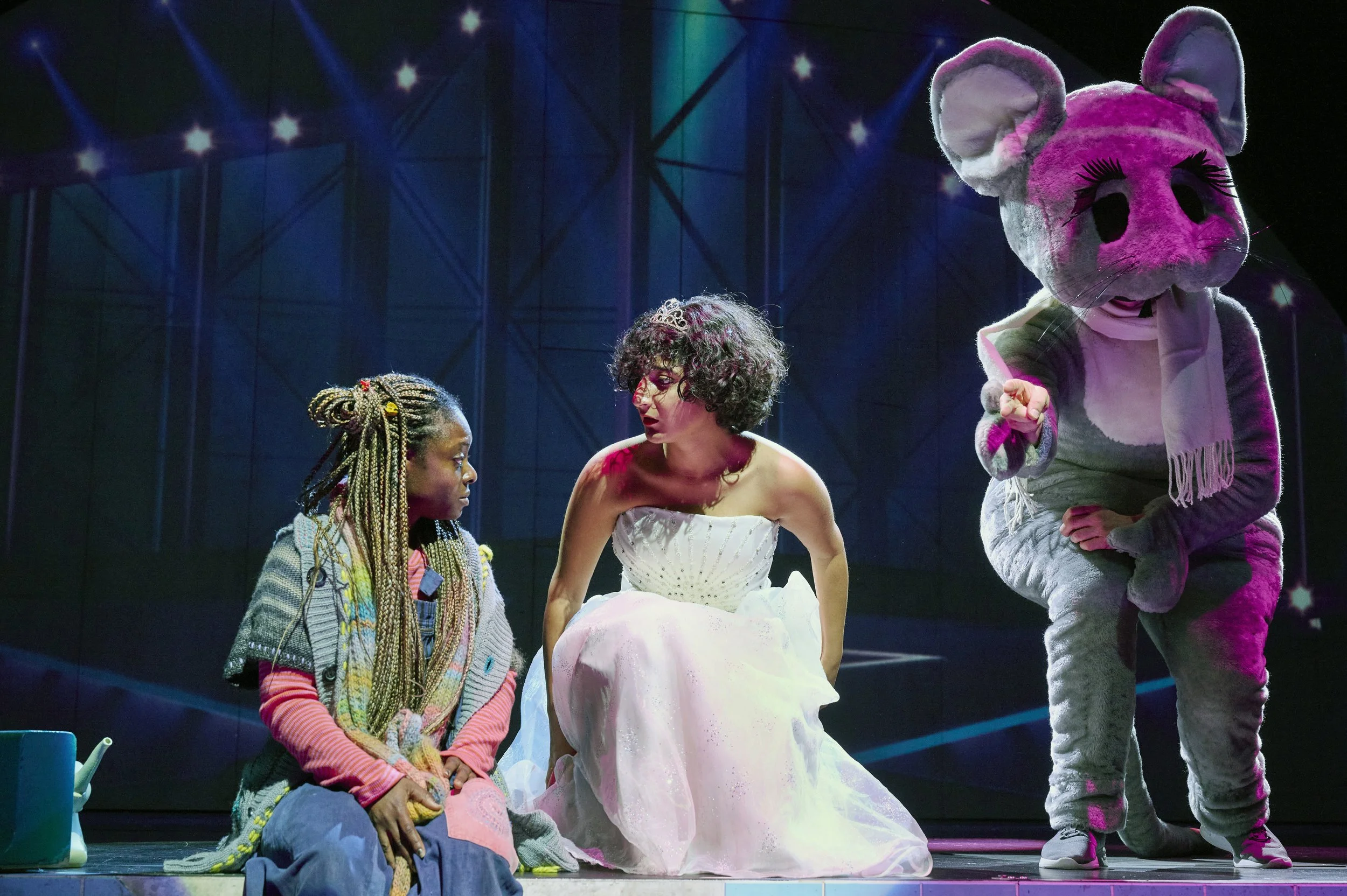 Drei Menschen auf einer Bühne, ein Mädchen mit geflochtenen Haaren, eine Frau im weißen Kleid und eine Person im Kuscheltier-Maus-Kostüm, während einer spricht und die anderen zuhören.