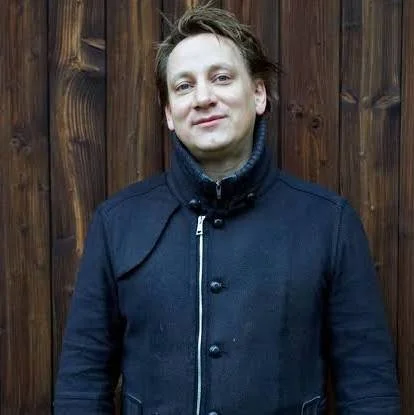 Eine Person mit kurzen, braunen Haaren trägt eine dunkle Jacke und steht vor einem Holzzaun.