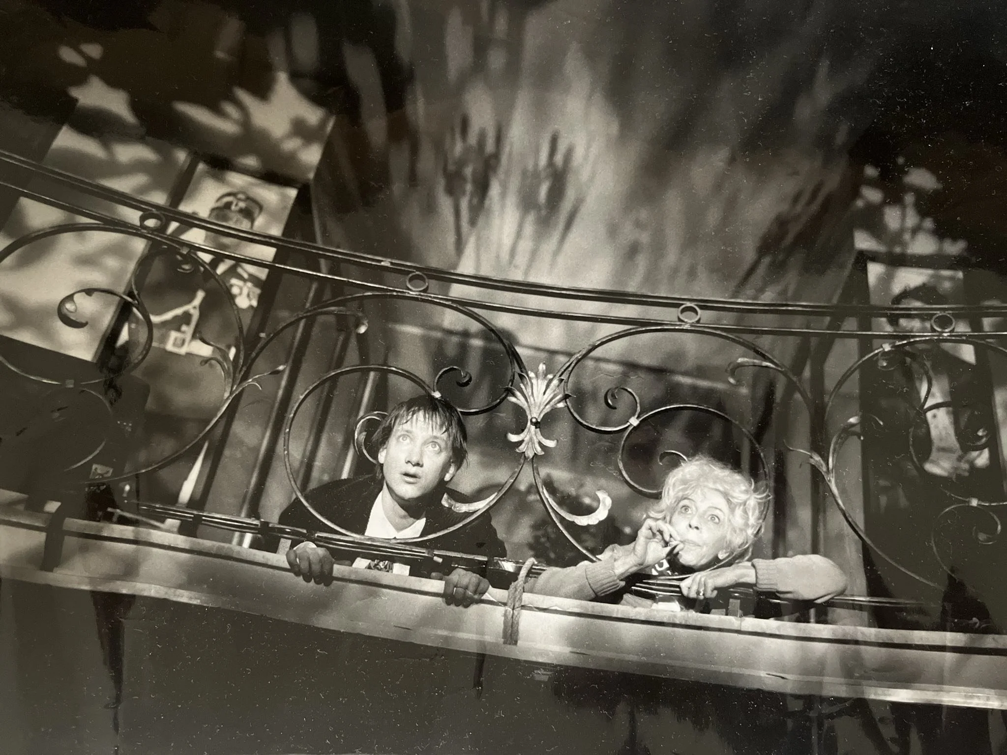 Zwei Kinder, ein Junge und ein Mädchen, schauen neugierig durch eine verzierte Metallabsperrung. Das Mädchen raucht eine Zigarette, während der Junge mit offenem Mund und weiten Augen schaut.