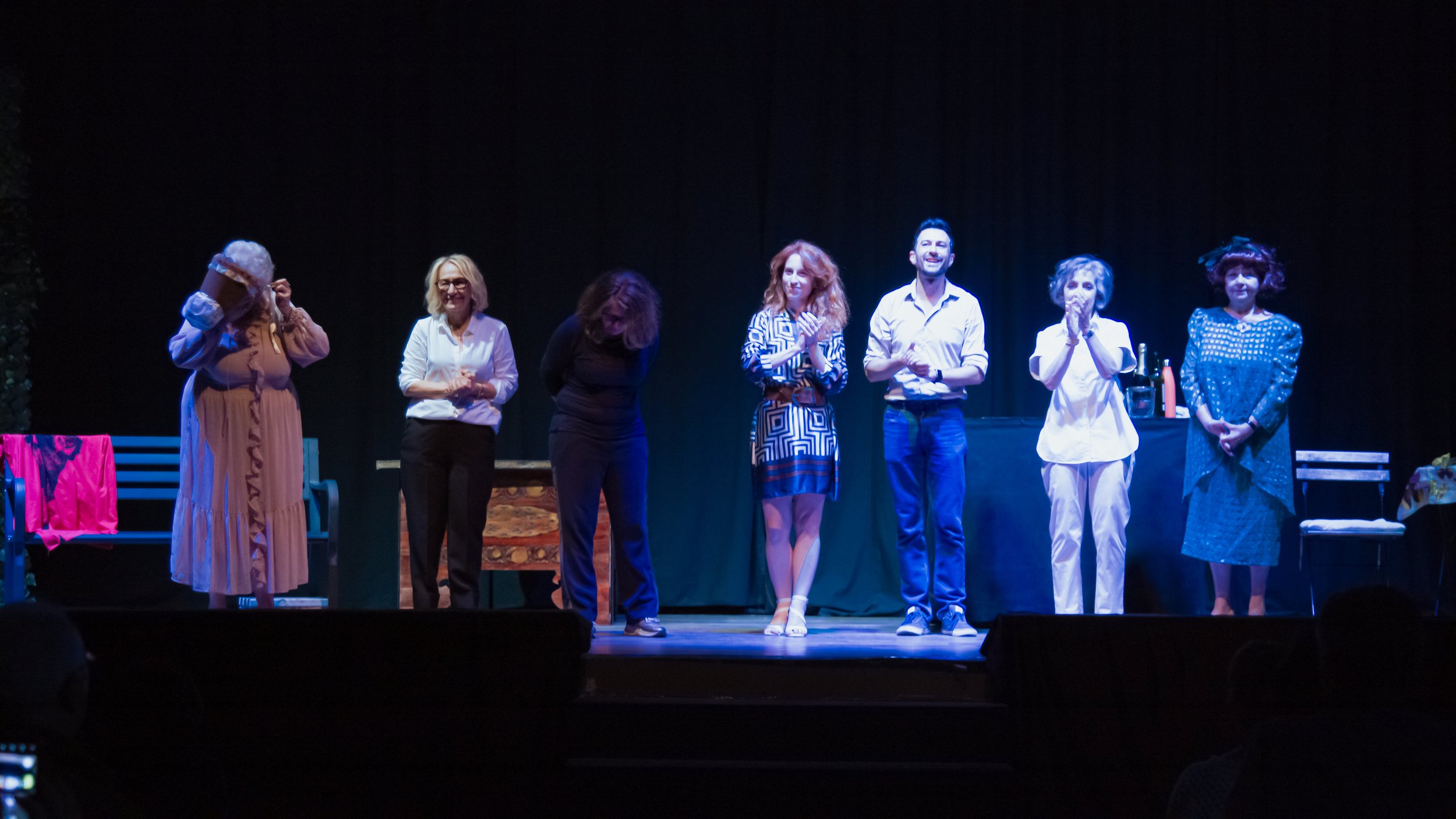 Un gruppo di persone si inchina sulla scena di un teatro, celebrando o salutando il pubblico, con tutti che sorridono, in un ambiente con sfondo nero.